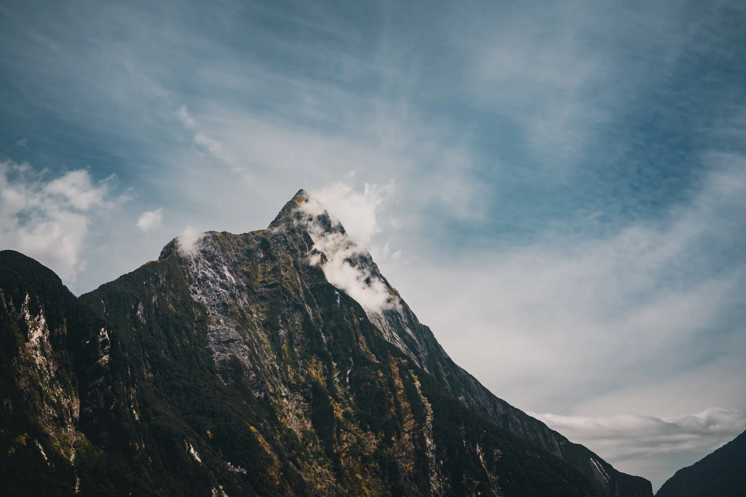 A large mountain with a rugged, green and rocky surface, partially covered by clouds, against a partly cloudy sky.