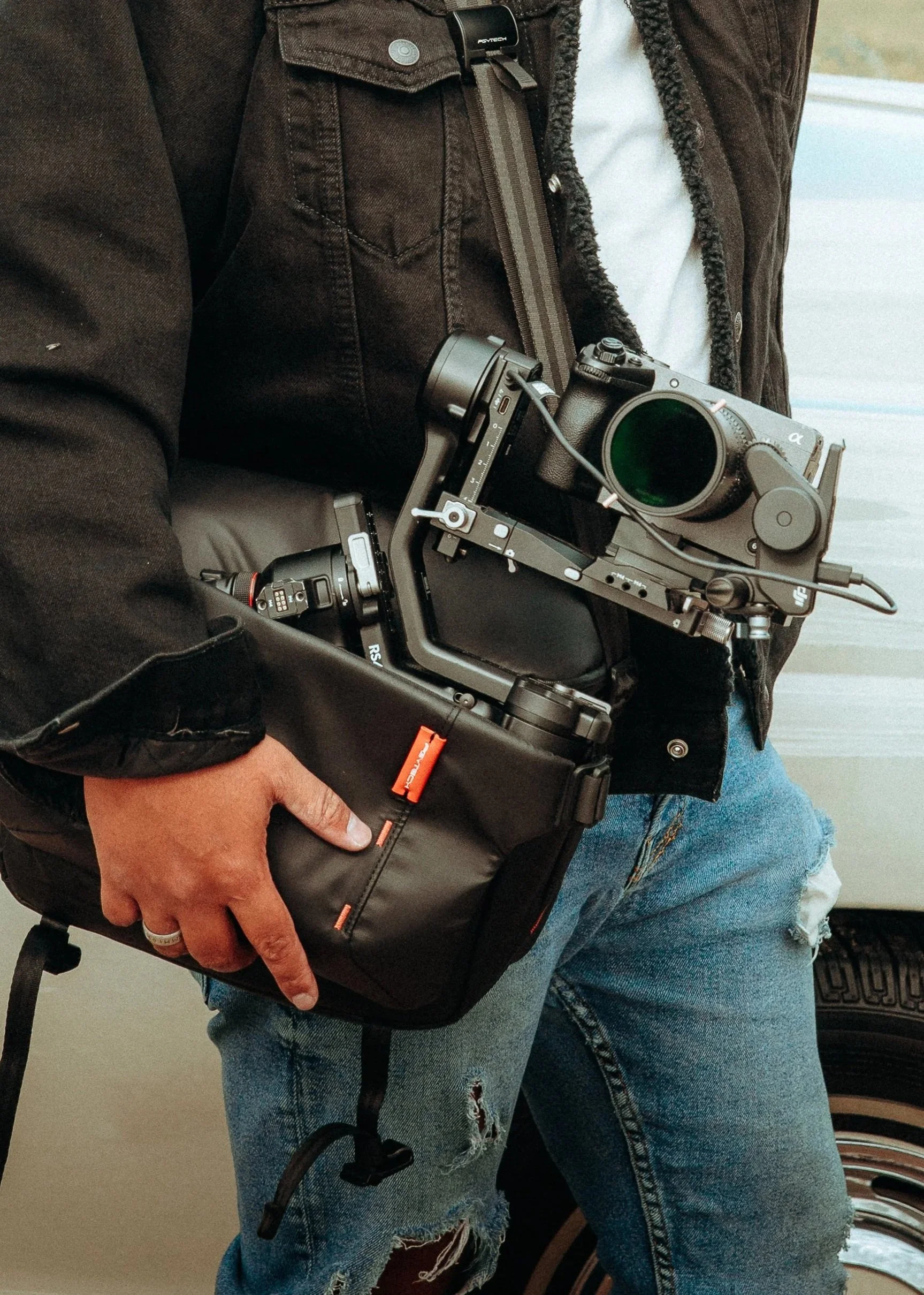 Brand Photography | Person wearing a black jacket and blue ripped jeans, holding a Sony Fx professional camera mounted on a DJI Ronin Gimbal and a small PGYTECH black bag.
