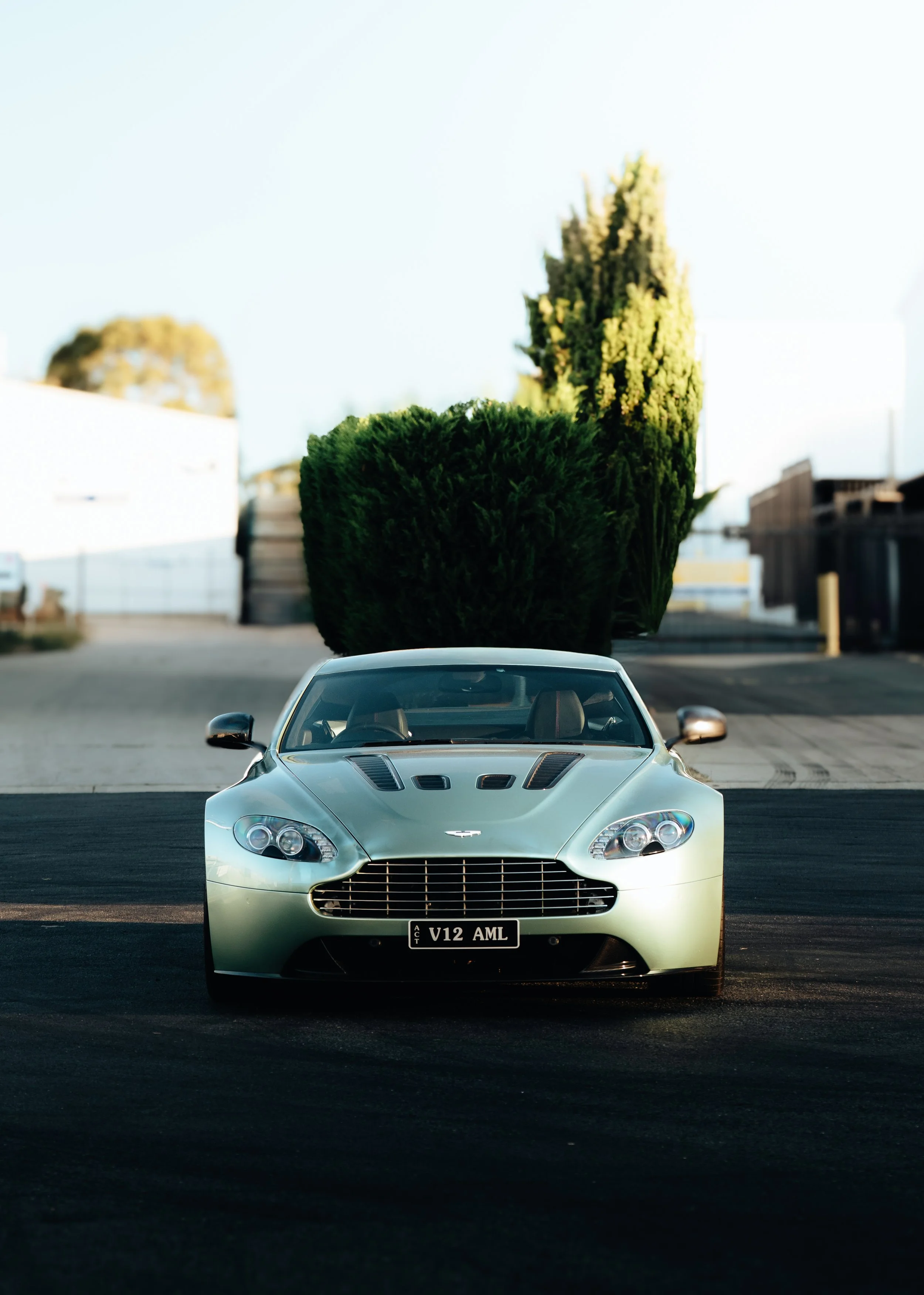 A silver Aston Martin V12 AML sports car parked in front of a large green hedgerow on a paved surface with an industrial background.