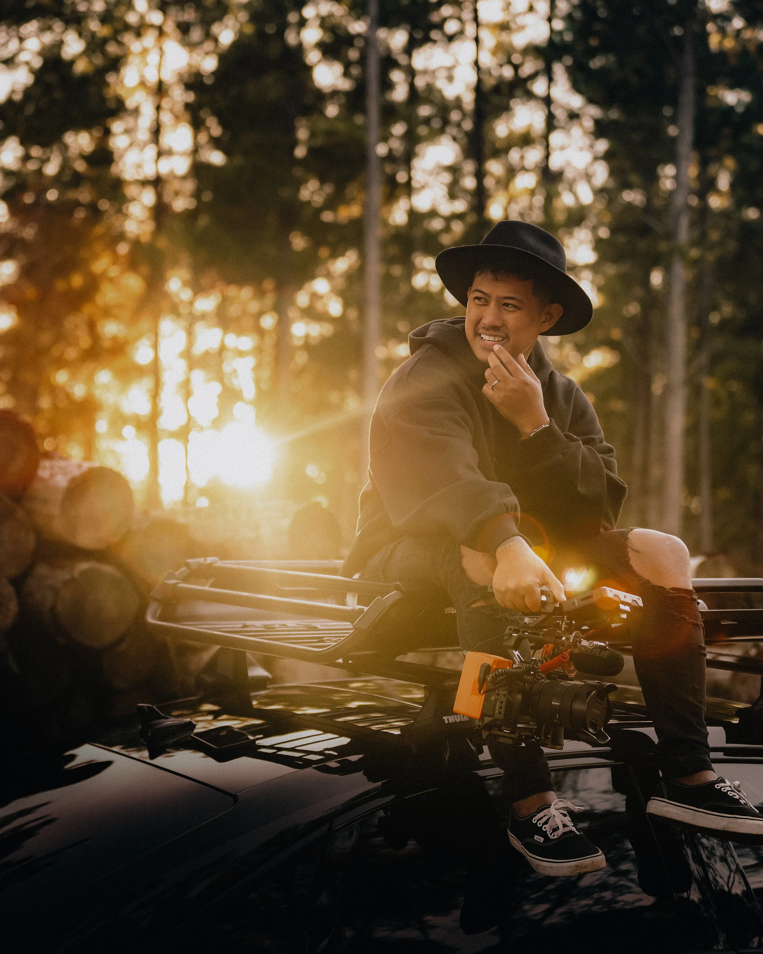 Jomel Montaos - Photographer and Videographer, Content Creator, sunrise posing - wearing a black wide-brim hat, black hoodie, ripped jeans, and black sneakers, surrounded by a forested area during sunset.