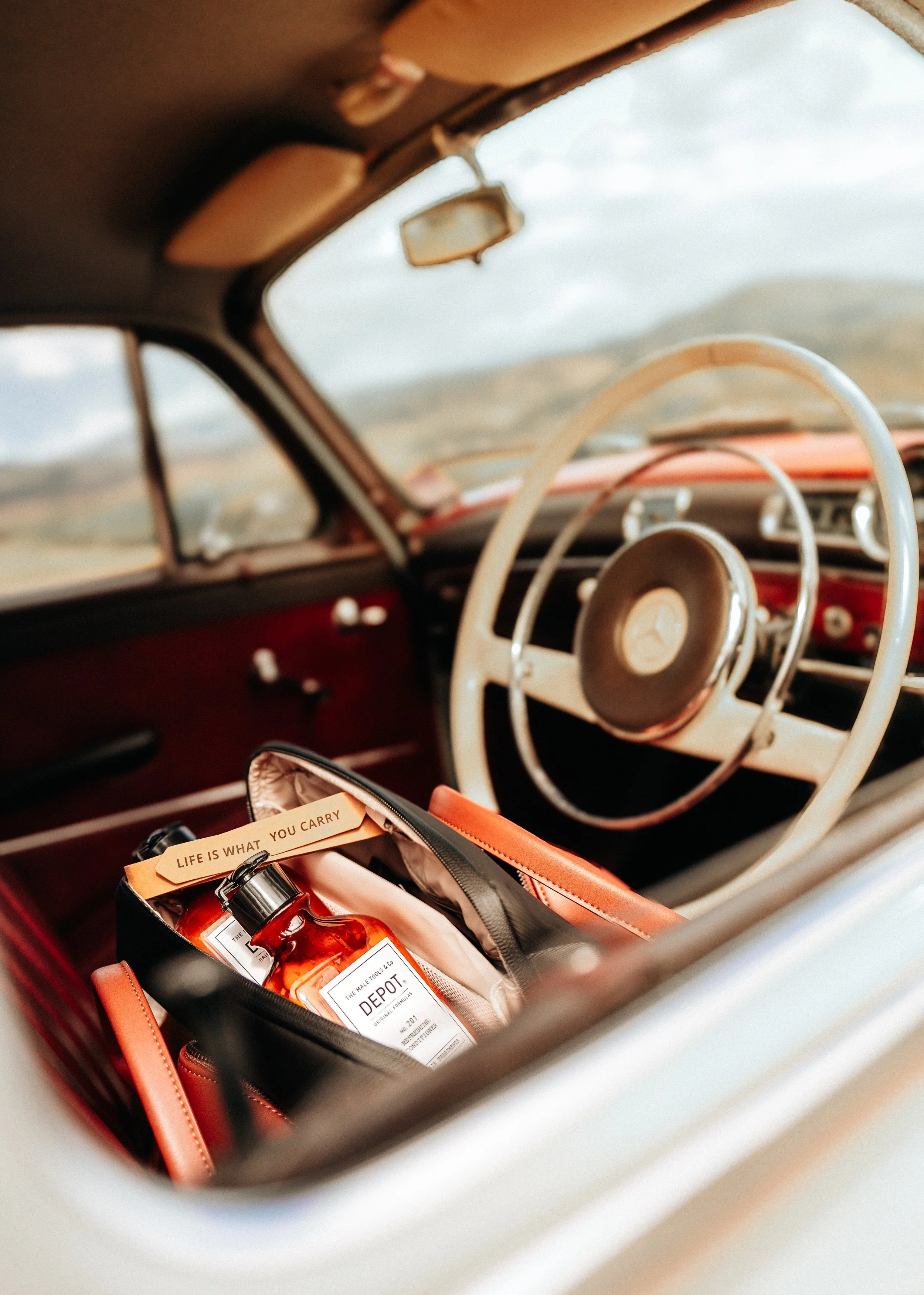 Brand Photography | Inside a vintage Mercedes with a red interior, view through the windshield showing a cinematic landscape, containing a bag with a brand labeled 'Depot' and a tag that reads 'Life is what you carry'.