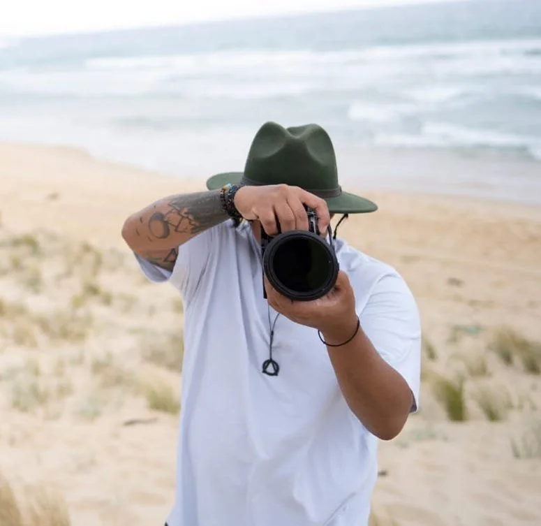 Person taking a photo with a professional camera on a beach, wearing a green wide-brimmed hat and a white t-shirt.