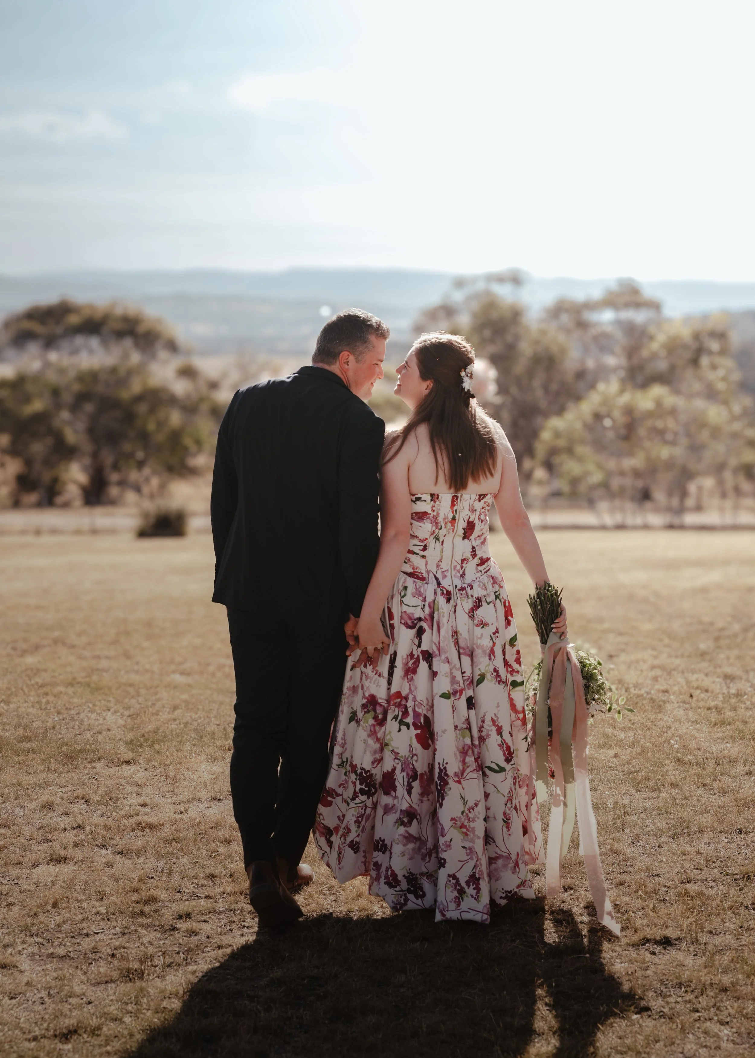 Wedding Photography |A couple standing face to face holding hands in an outdoor field during daylight, with the woman holding a bouquet of flowers and wearing a floral dress, and the man in a dark suit, with trees and distant hills in the background.