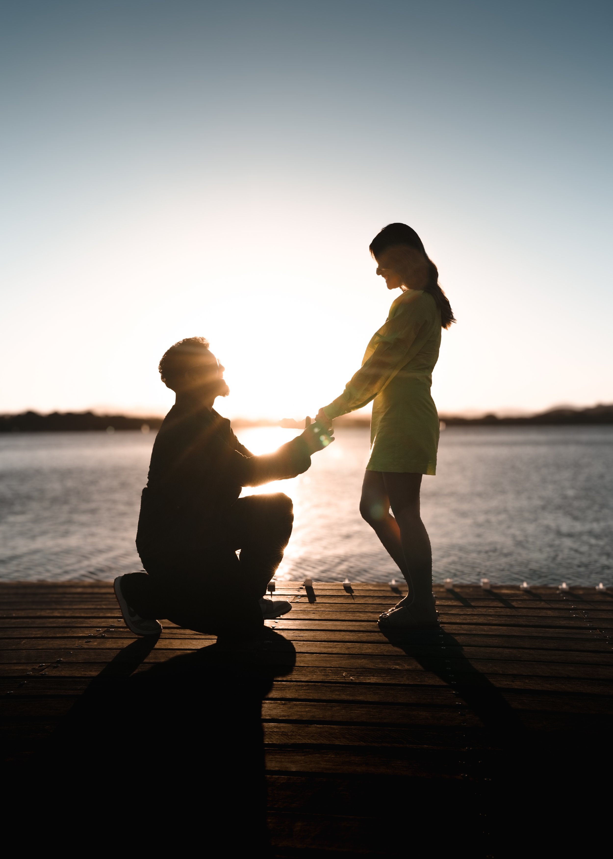 Engagement Photography | A silhouette of a man kneeling and proposing to a woman on a dock near the water during sunset. Silhouette, Sunset, proposal and engagement photography.