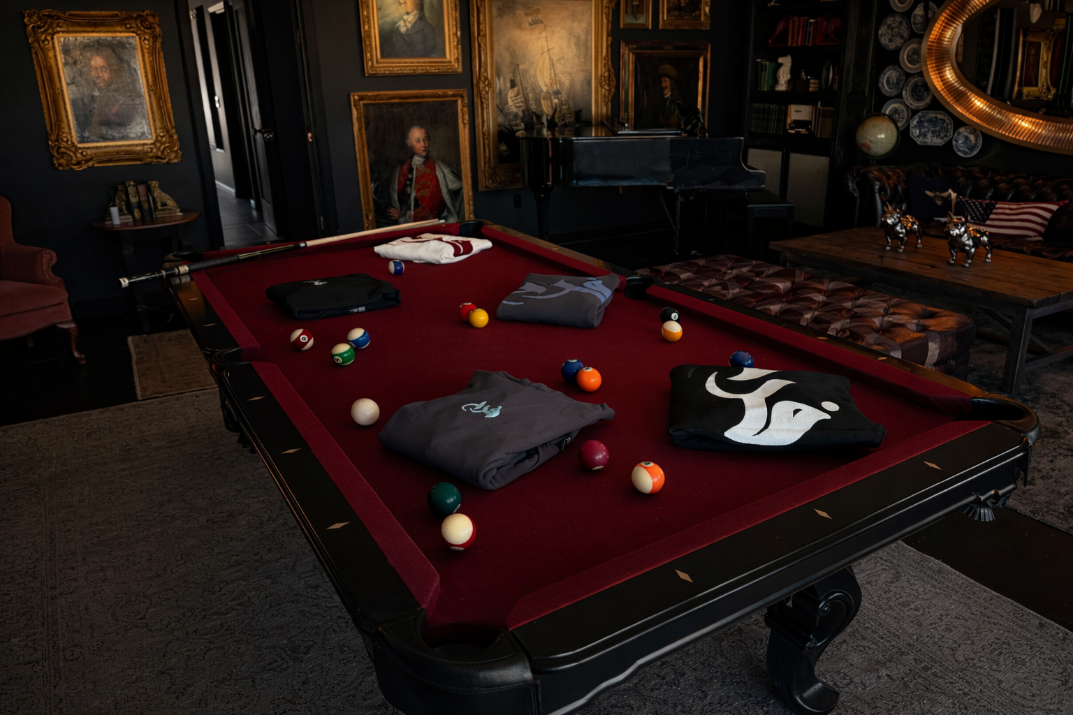 A billiards table with pool balls, pool cues, and folded clothes in a room decorated with framed paintings, a grand piano, a mirror, and decorative plates.