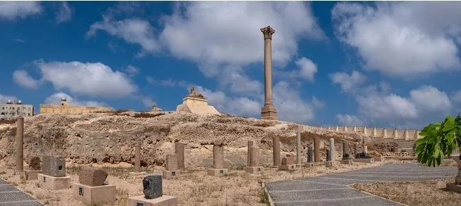 Ancient ruins with stone columns and a tall Roman-style column under a cloudy sky.