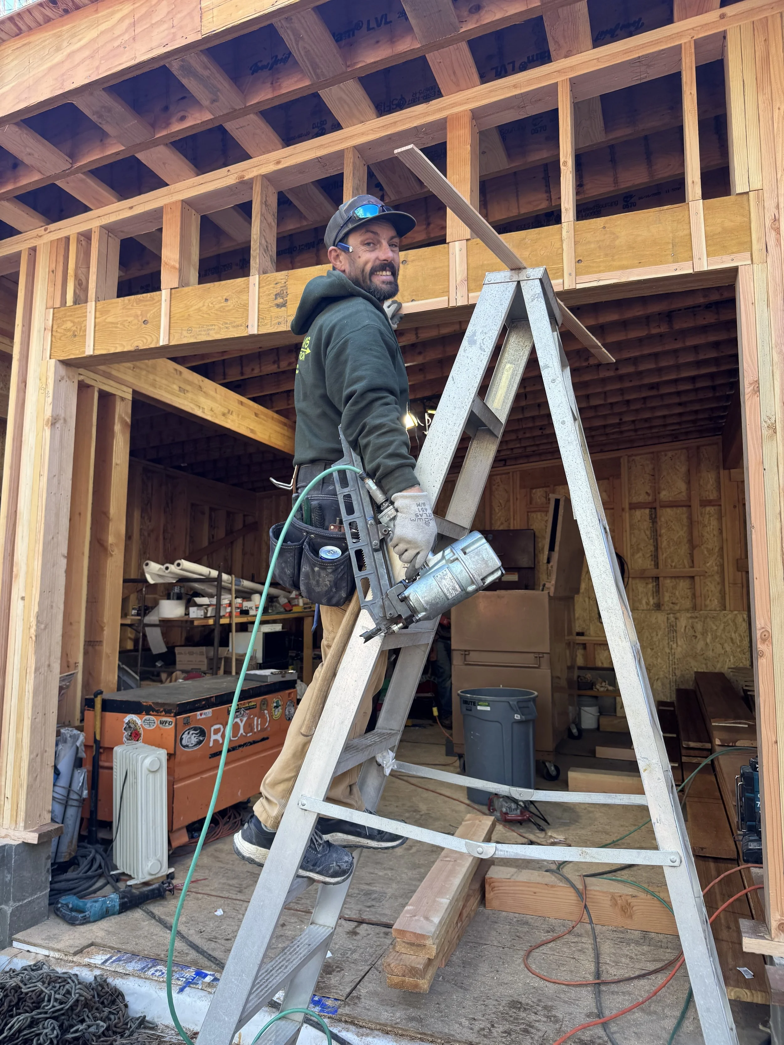 A construction worker standing on a ladder at a building site, holding a nail gun, with the wooden framing of a house under construction in the background.