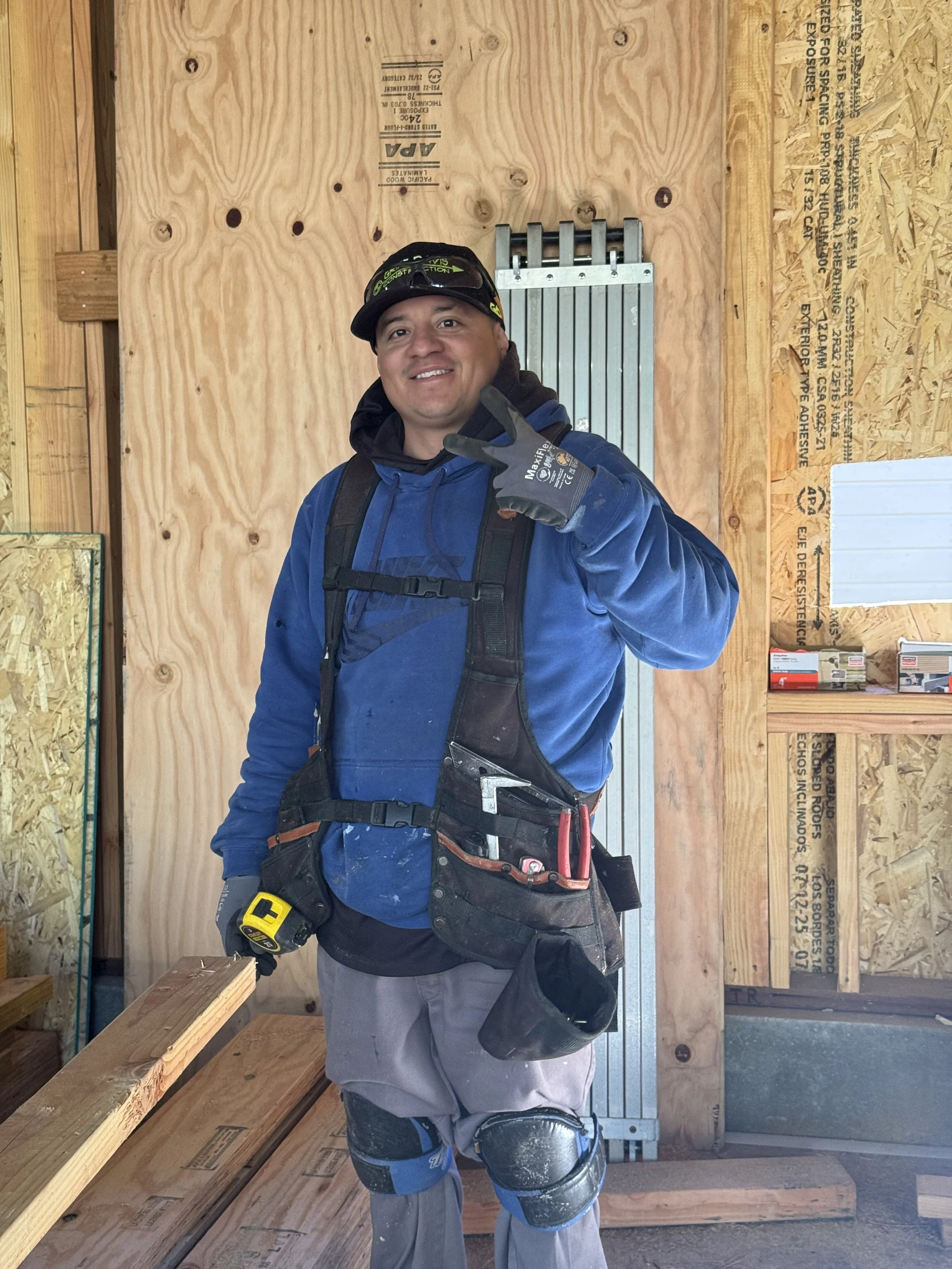 A smiling construction worker wearing a blue jacket, work gloves, and knee pads, standing inside a wooden building under construction, making a peace sign with his hand.