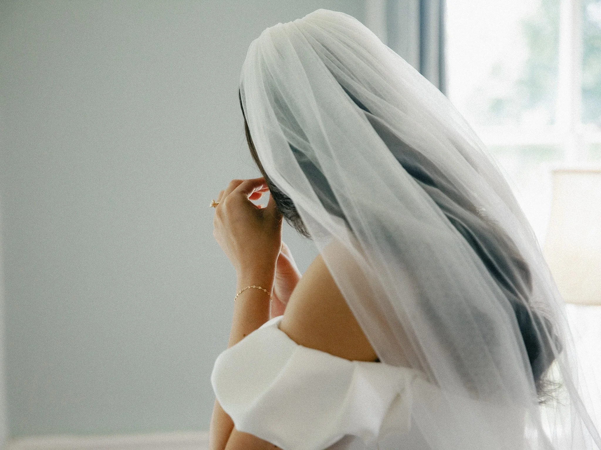 Bride in an off-shoulder dress adjusting her long veil by the window