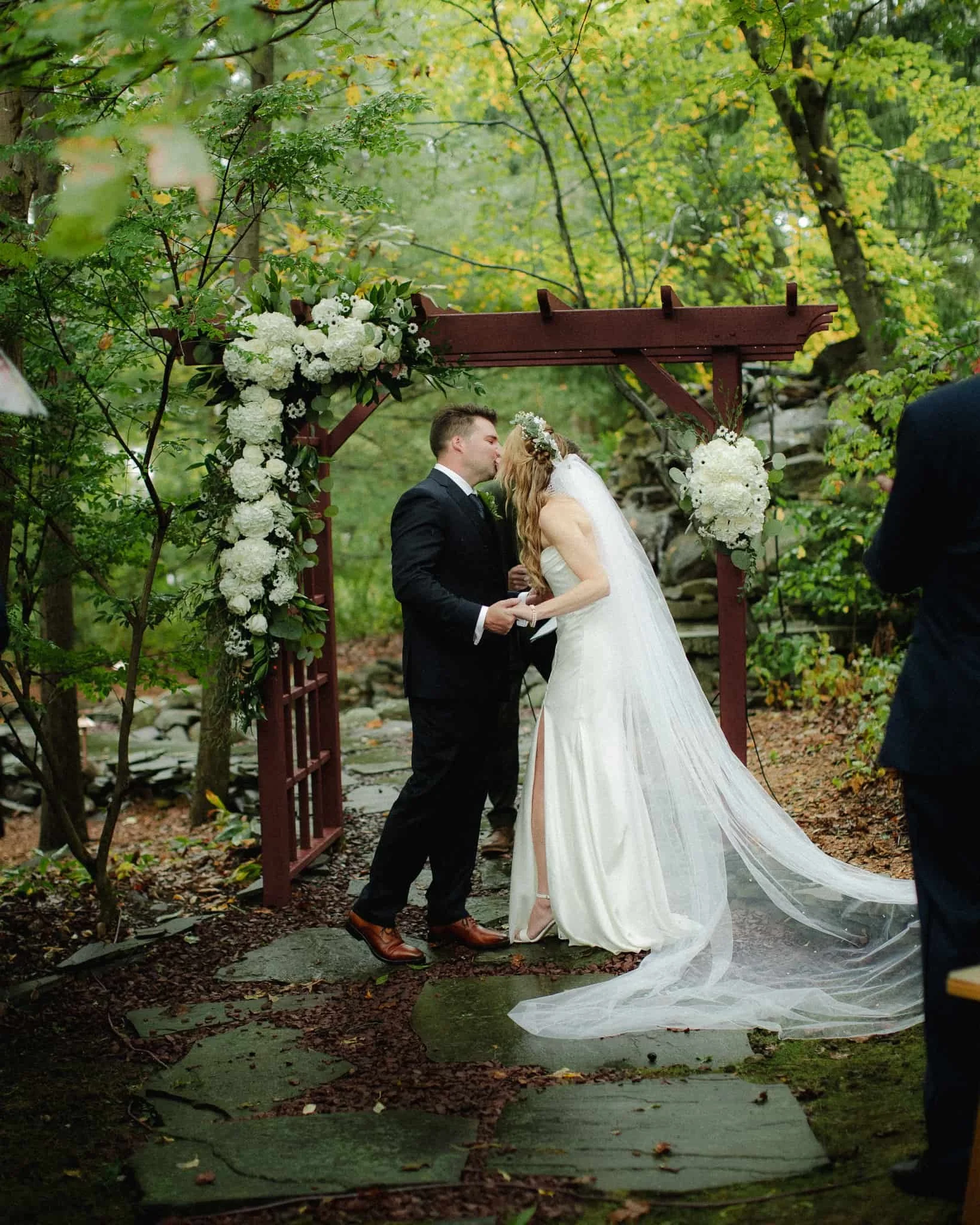 Bride and groom kissing beneath a floral wooden arbor in a wooded garden