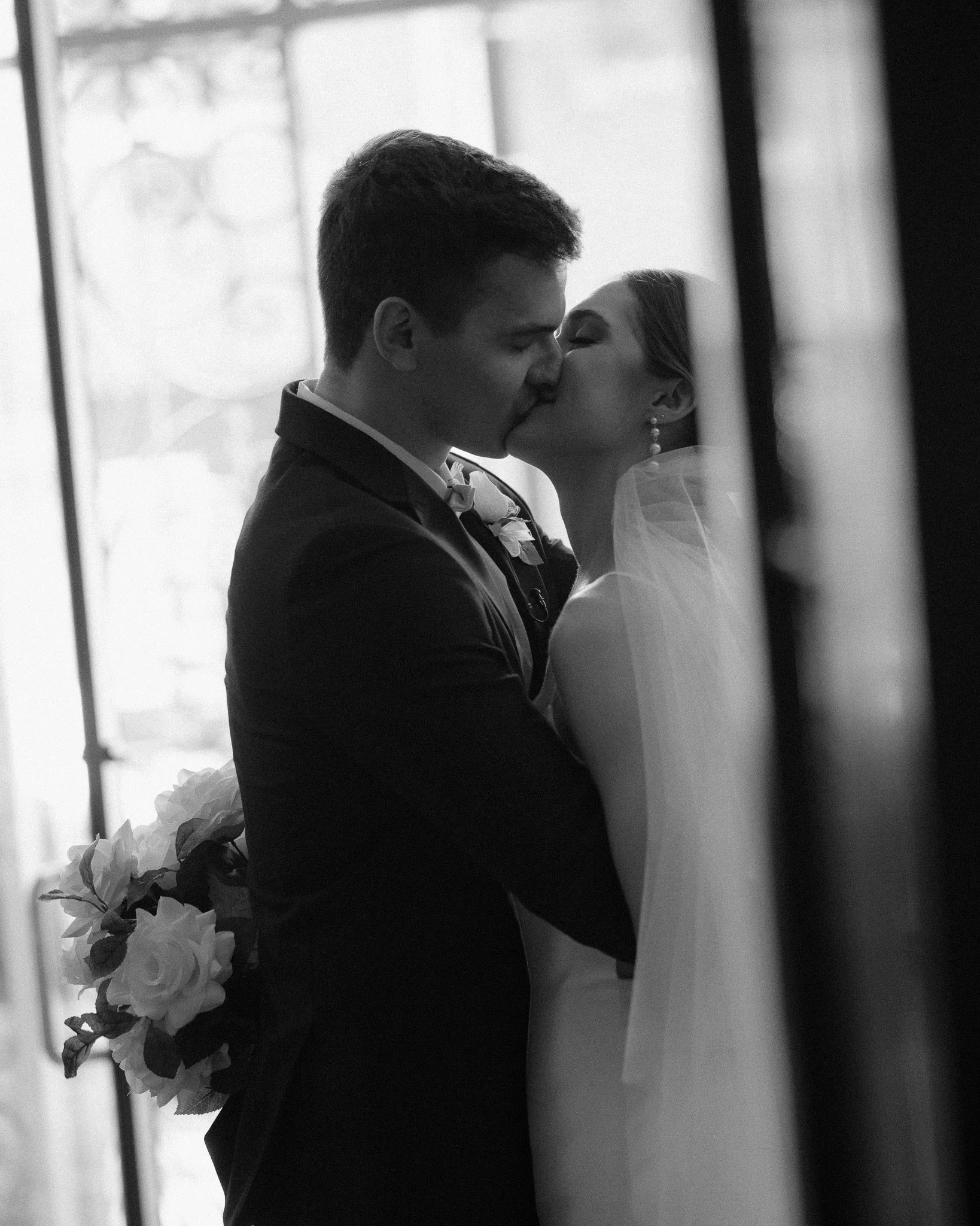 Black and white portrait of bride and groom kissing by a window