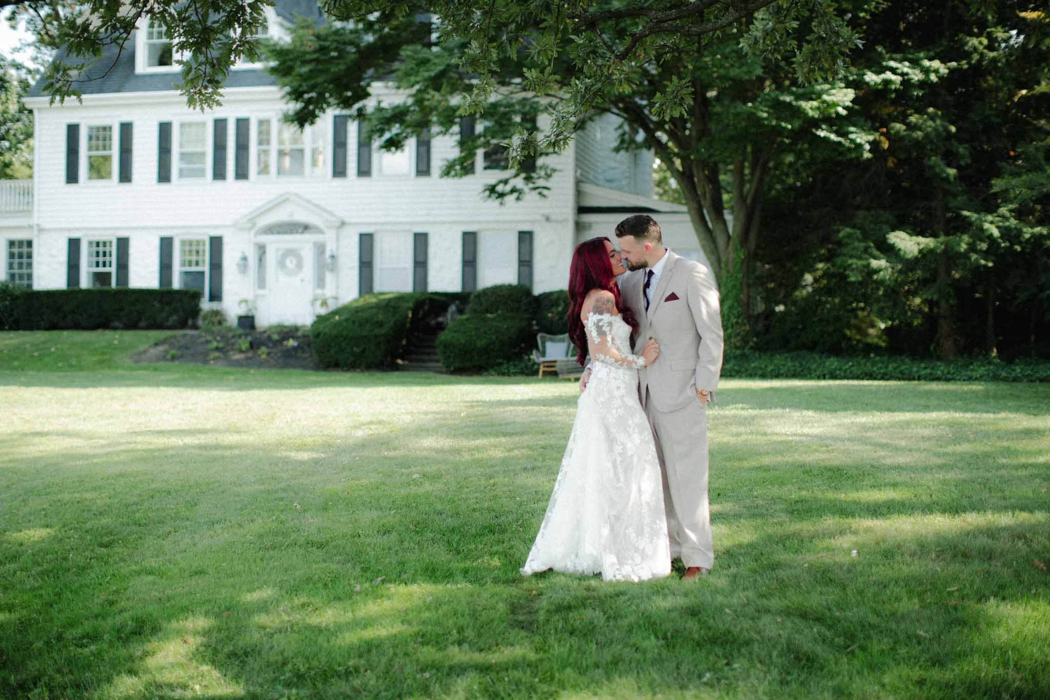 Bride and groom kissing on the lawn in front of a white colonial house