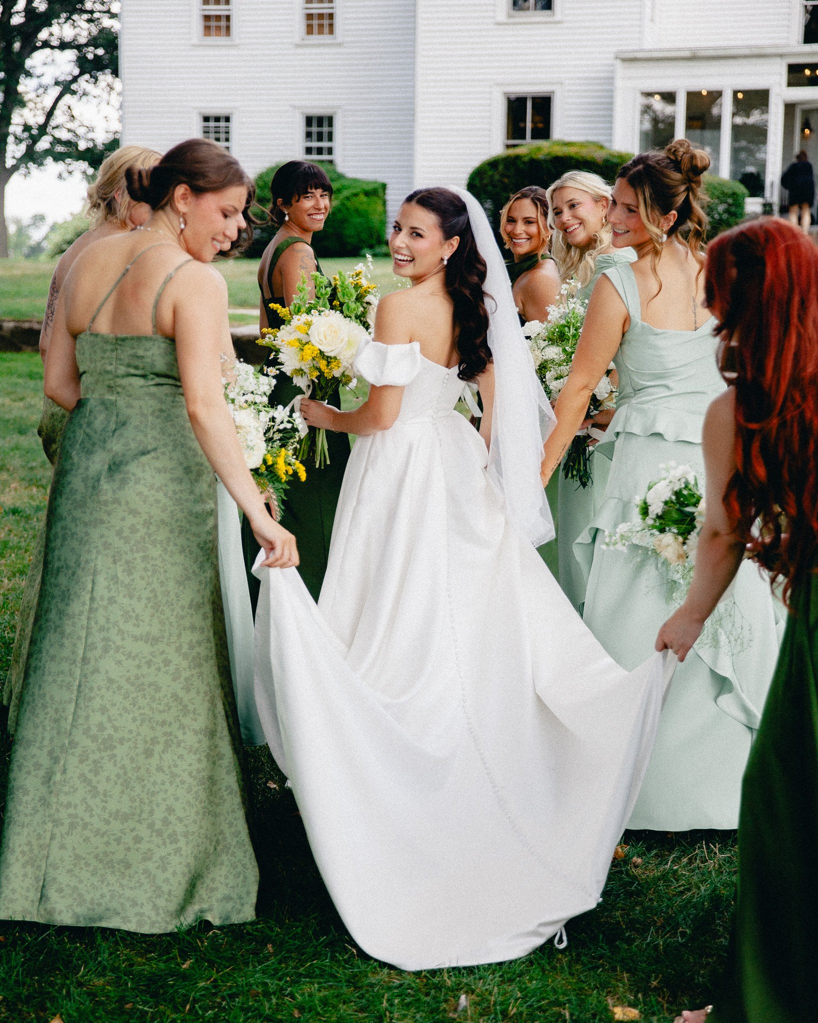 Bridesmaids in green helping the bride with her gown on the lawn