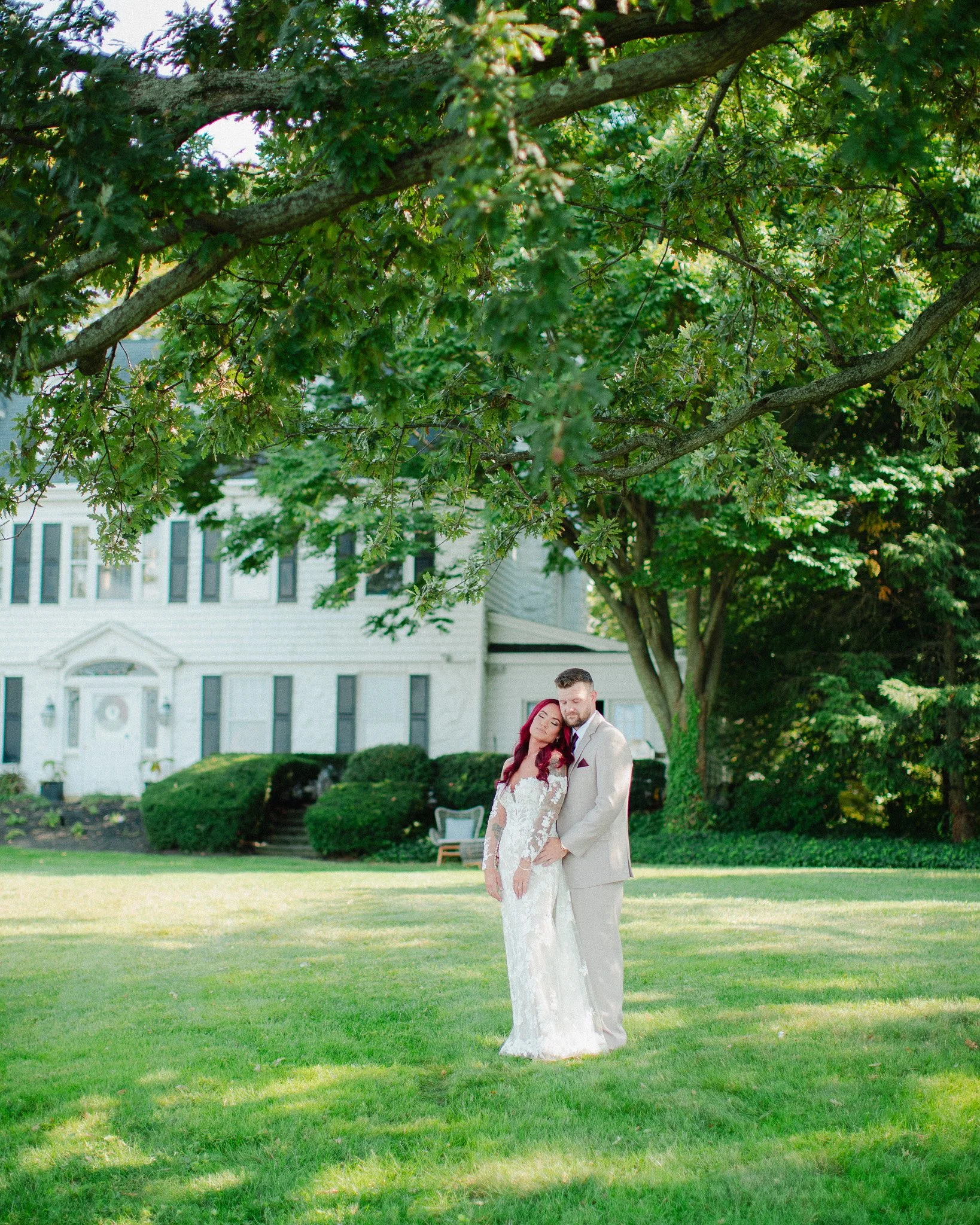 Bride and groom on the lawn beneath a large tree by a white colonial house