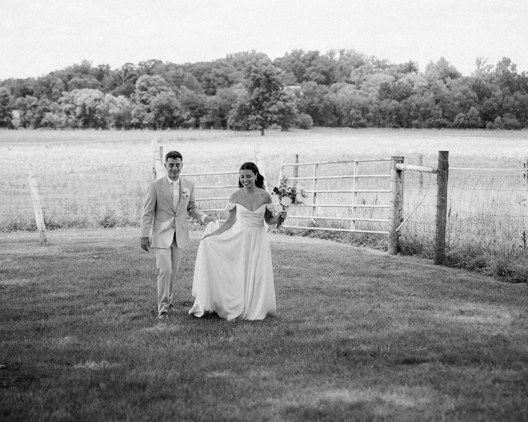 Bride and groom walking through a farm field by a wooden fence, black and white