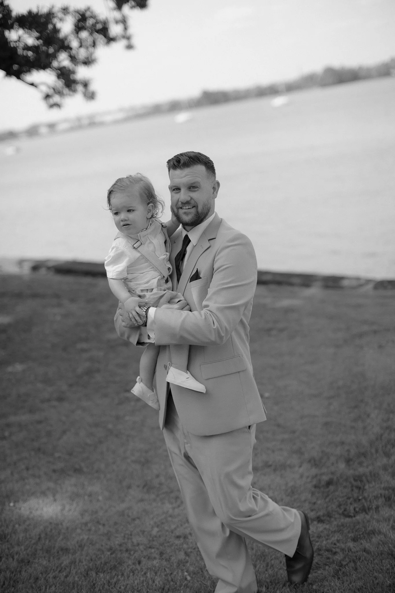 Groom holding a smiling toddler by the water, black and white wedding photo