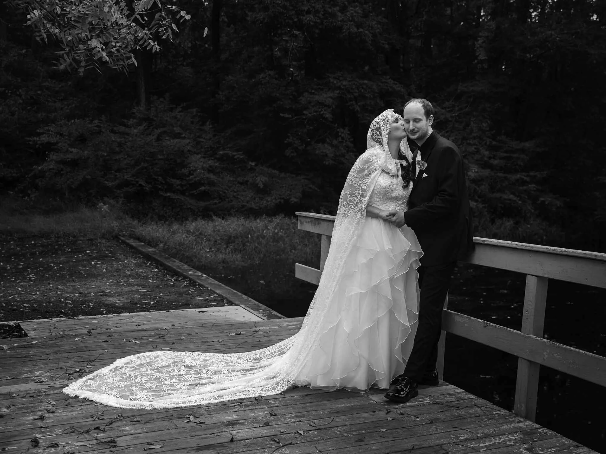 Bride and groom on a wooden footbridge, long veil trailing, black and white