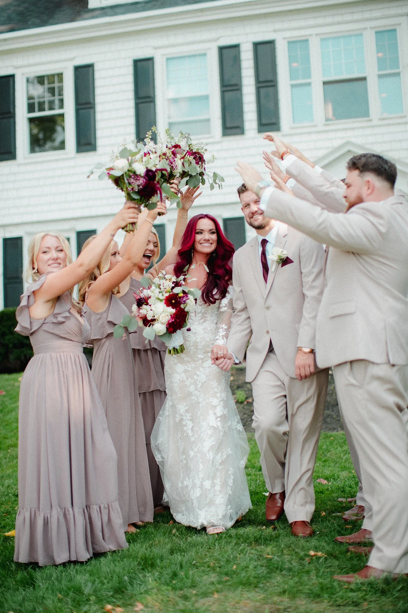Bridal party cheering around bride and groom outside a white colonial house