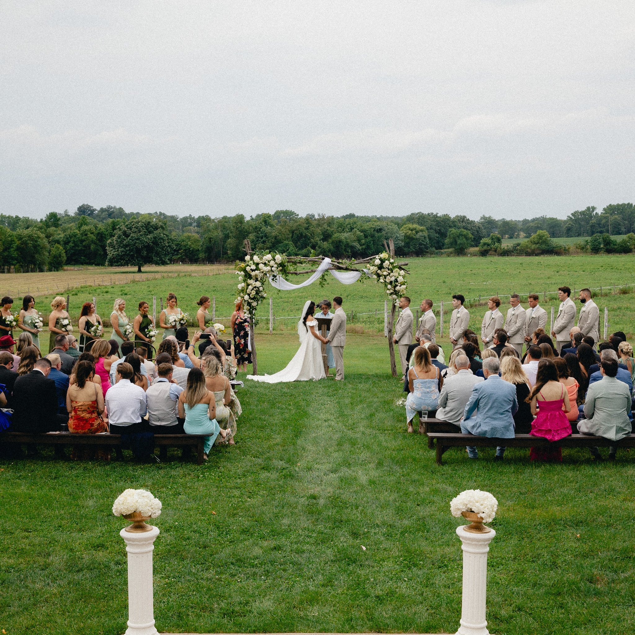 Outdoor wedding ceremony on a grassy field with guests and floral arch