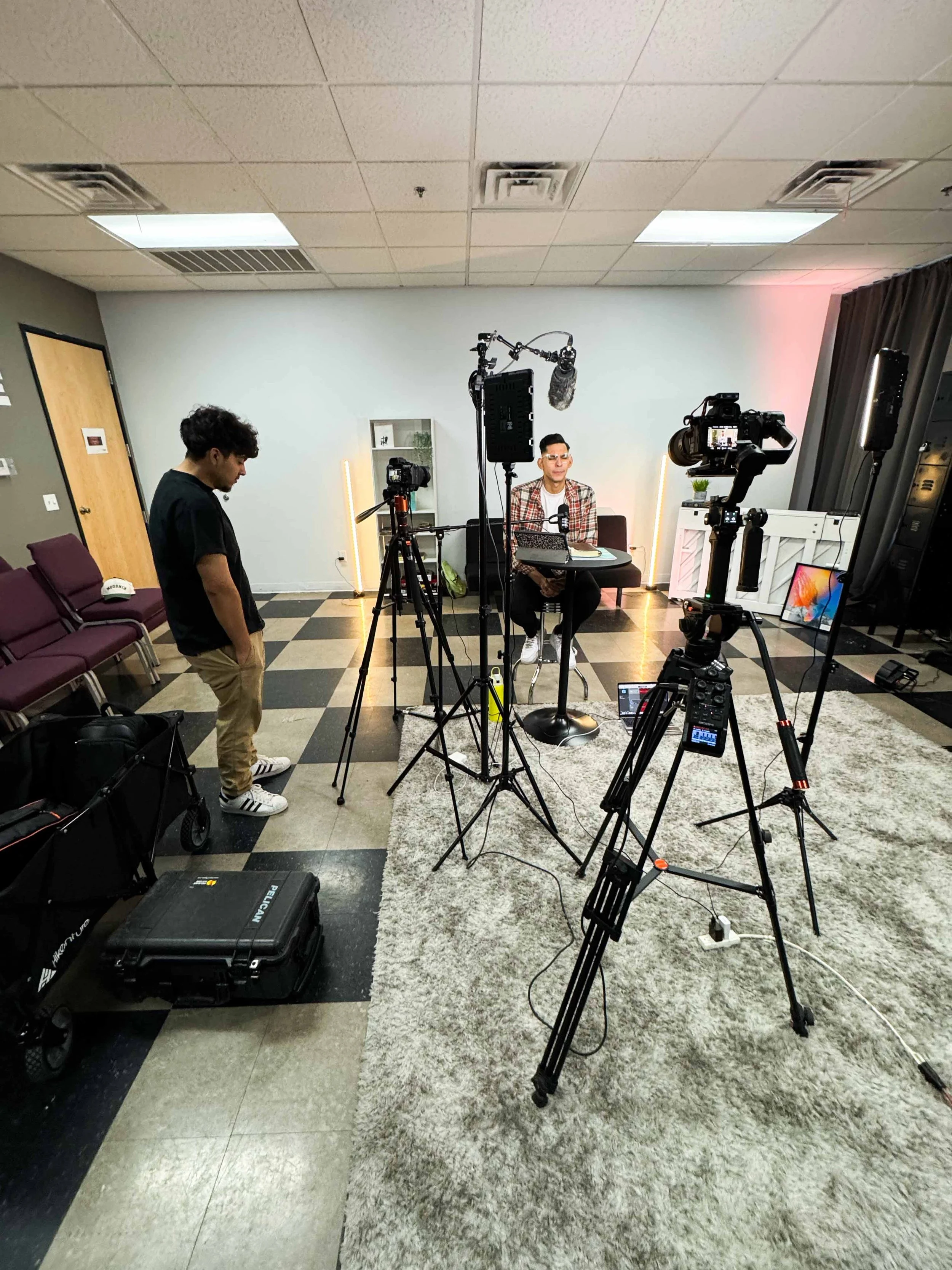 A video interview or recording setup with cameras, lighting, and a person sitting at a table with a laptop and microphone, in a room with chairs and equipment.