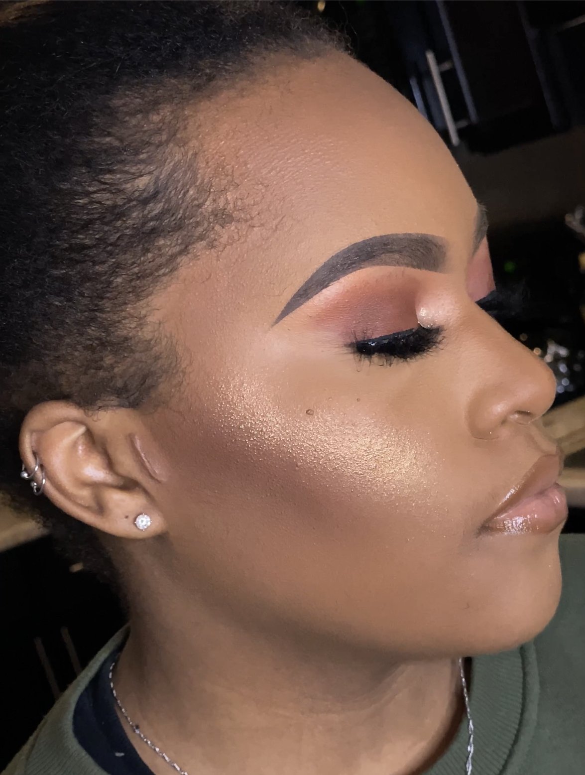 Close-up of a person with makeup featuring bold eyebrows, eye makeup, shimmering highlighter, and lip gloss.