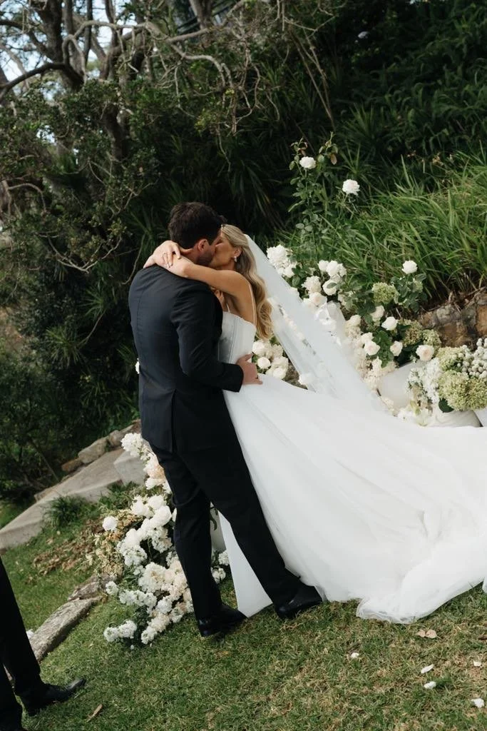 A bride and groom share a kiss outdoors during their wedding, with the bride in a white gown and the groom in a black suit, surrounded by white flowers and green foliage.