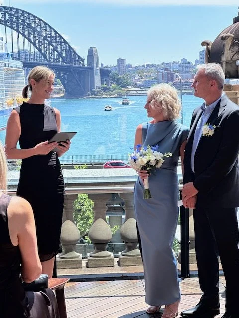 A wedding ceremony outdoors with the Sydney Harbour Bridge in the background. A woman in a blue dress holding a bouquet is standing next to a man in a suit, while a woman in a black dress appears to be officiating or speaking.