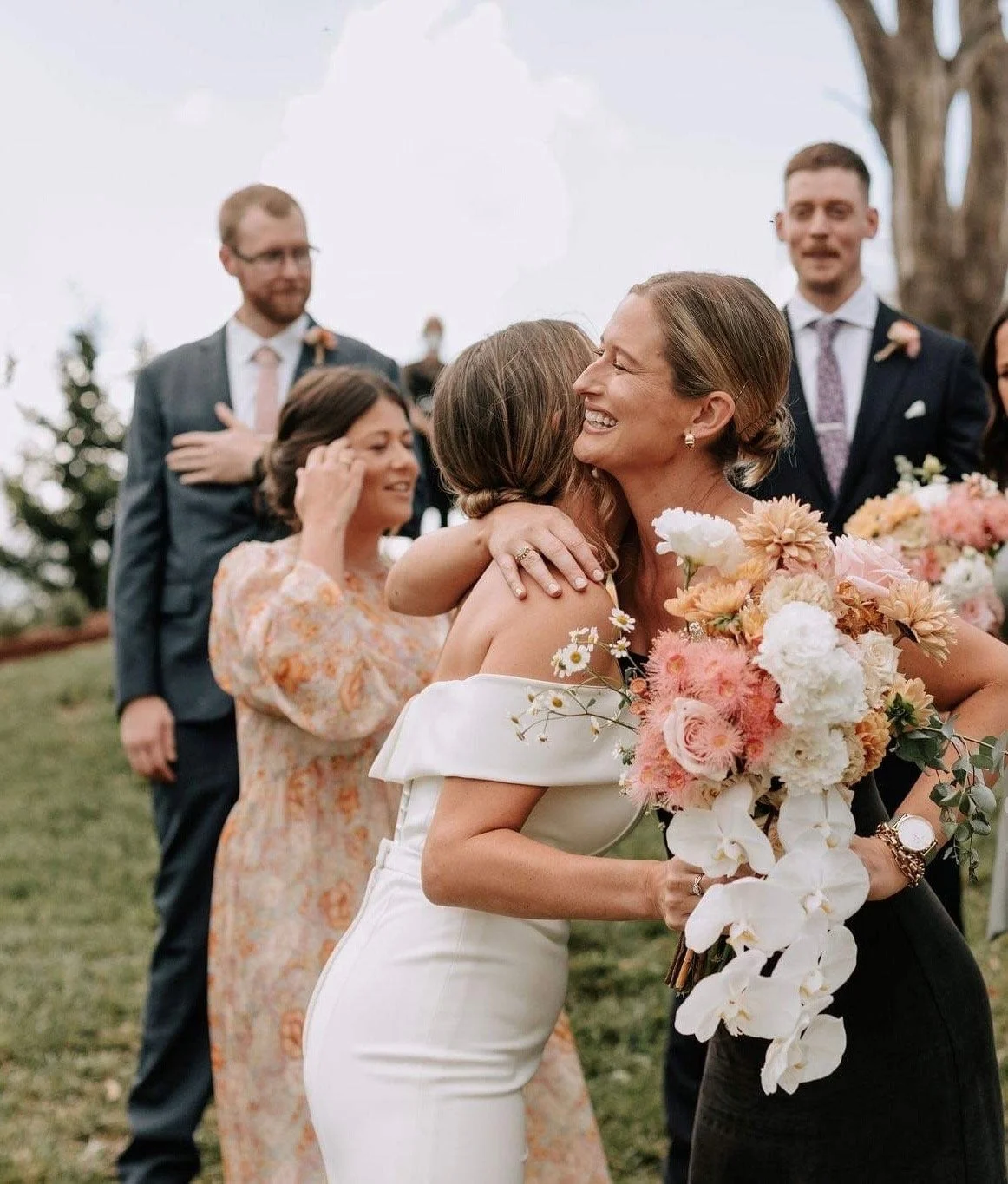 Two women hugging at a wedding, with one woman holding a large bouquet of flowers. In the background, other wedding guests are standing outdoors, smiling and celebrating.