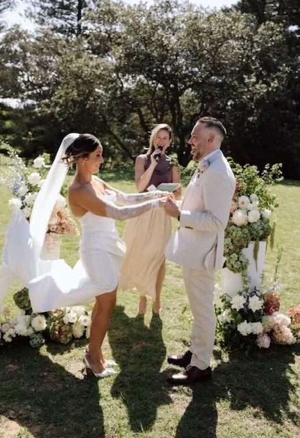 Bride and groom exchanging vows outdoors with a woman officiant and floral decorations.