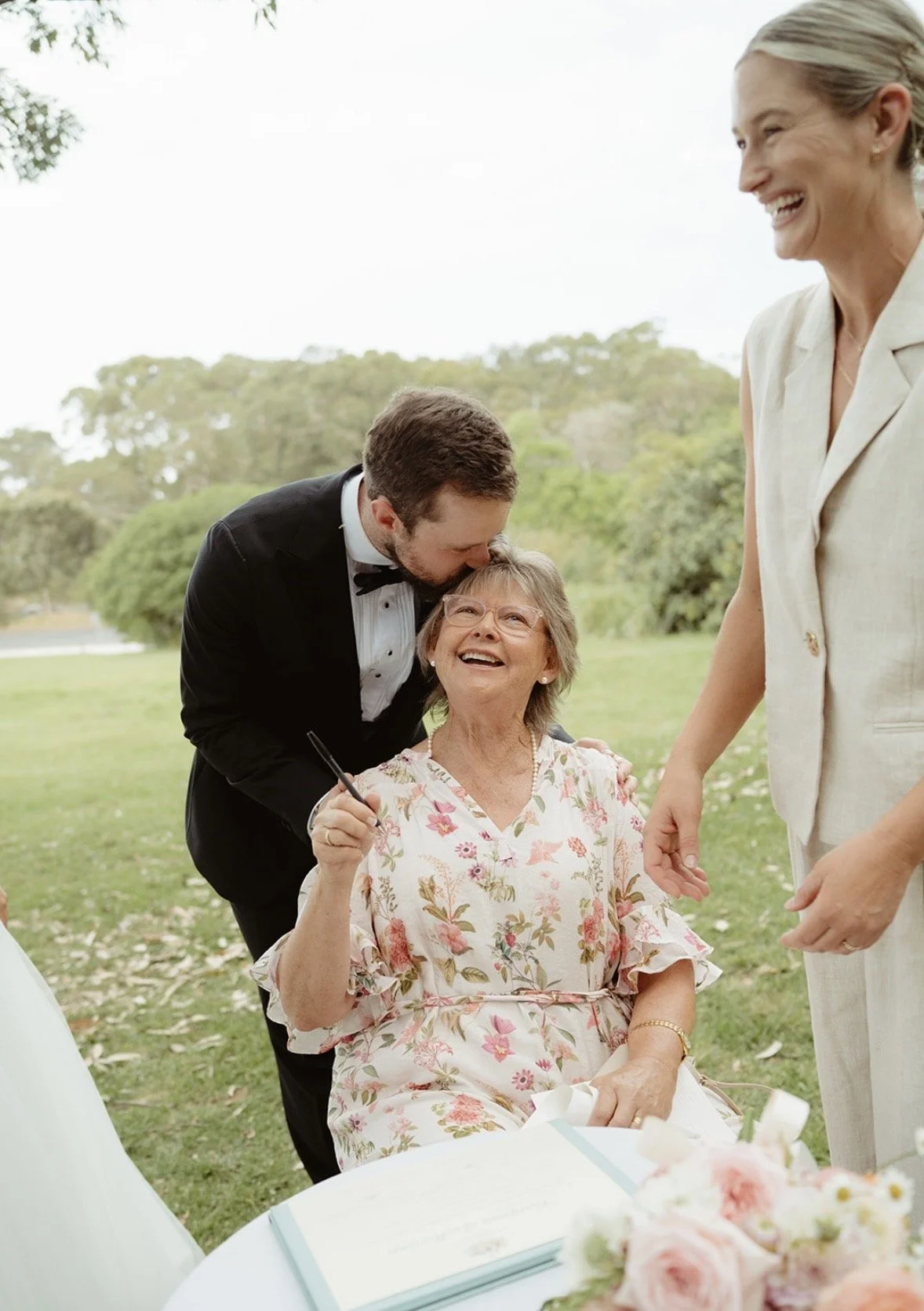A woman in a floral dress is seated outdoors, smiling as a man in a tuxedo kisses her forehead, while another woman stands next to her, smiling gently.