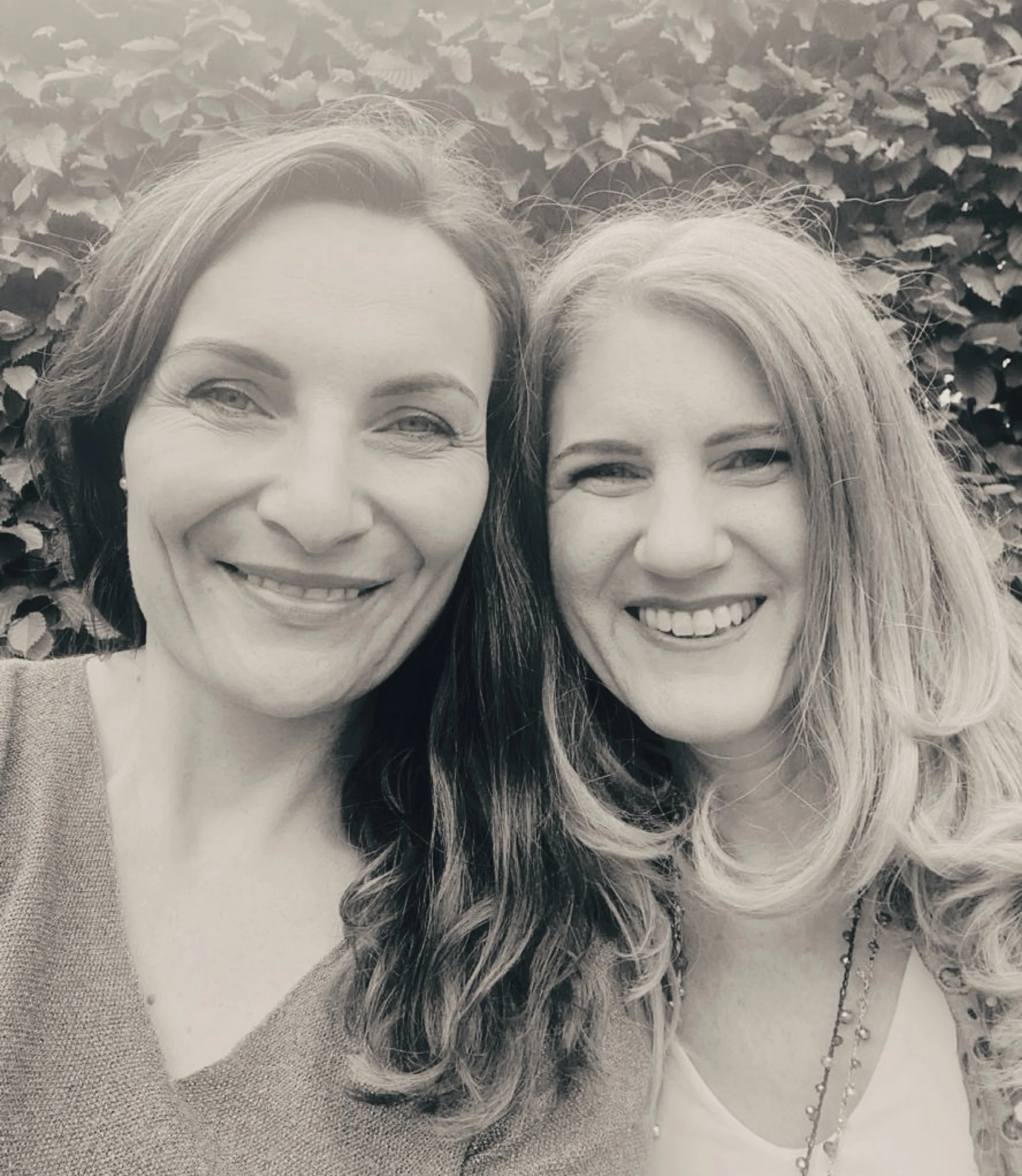 Zwei Frauen lächeln und nehmen ein Selfie vor einer Hecke auf.