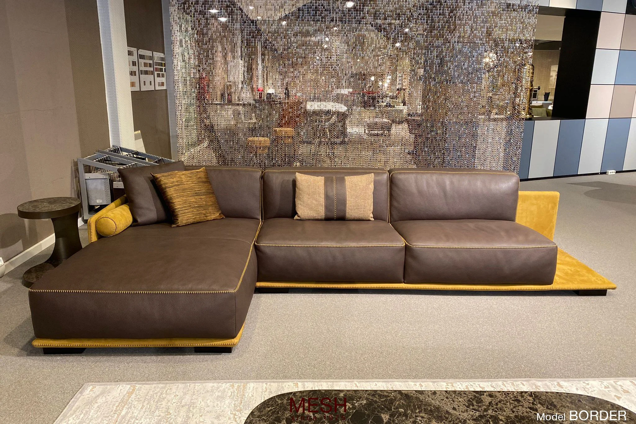 Een moderne zithoek met een grote leren hoekbank in bruin en geel, versierd met kussen. Achter de bank bevindt zich een decoratieve scheidingswand van spiegels en een open keuken met eetgedeelte.