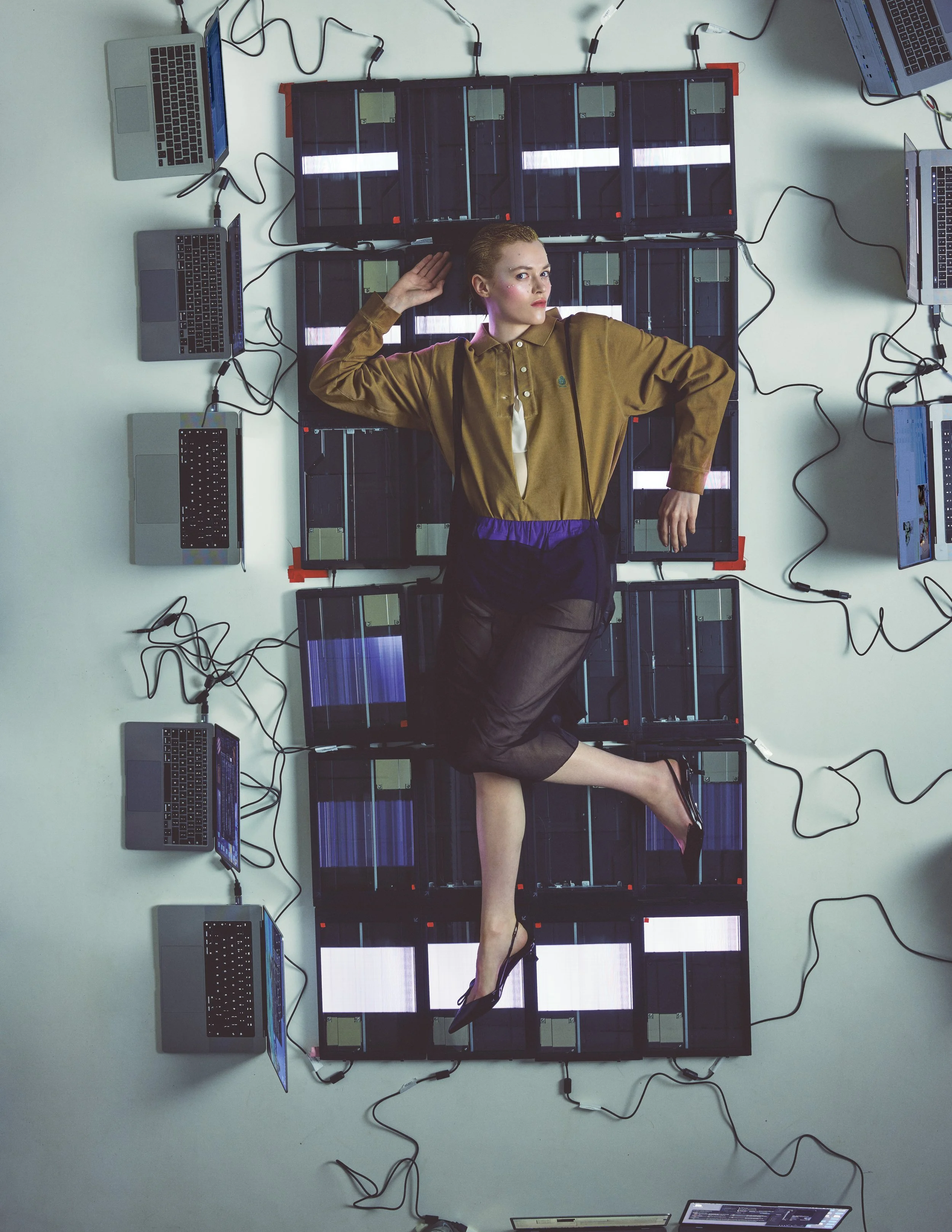 A woman lying on a white table, surrounded by multiple open laptops connected with black wires, with a black grid-like structure behind her. The outfit is from Prada. 