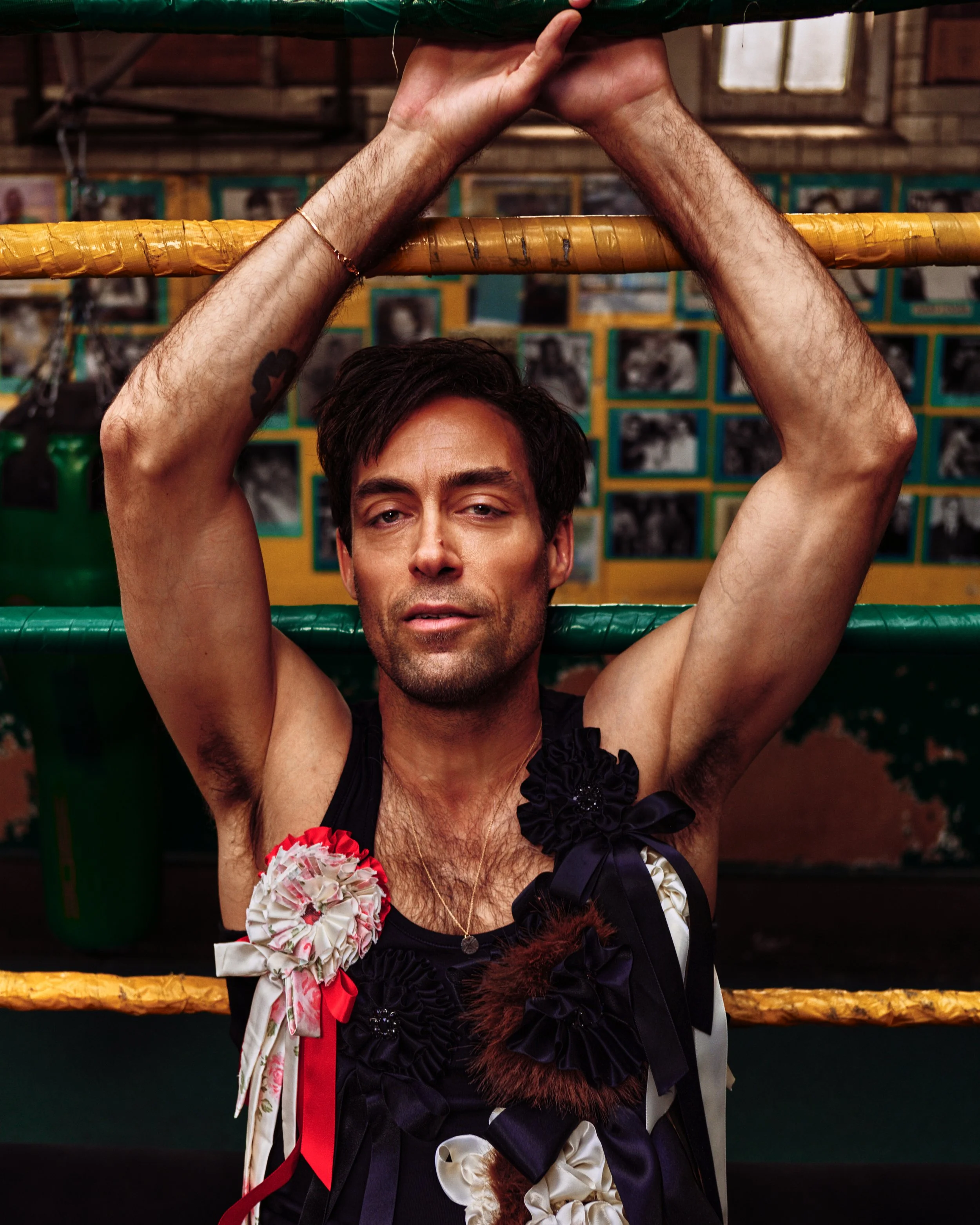 A man with dark hair and tan skin, wearing a black dress with floral and bow decorations, is holding onto a yellow gymnastic ring with both hands. He is standing in a gym with a wall of framed photographs in the background.