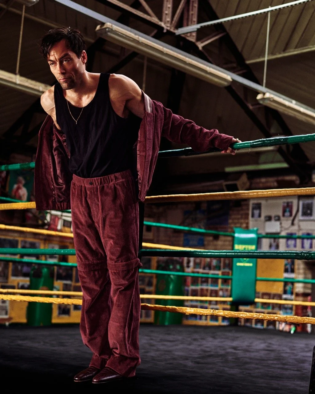A man in a black tank top and maroon corduroy pants leaning on a boxing ring's ropes in a gym.