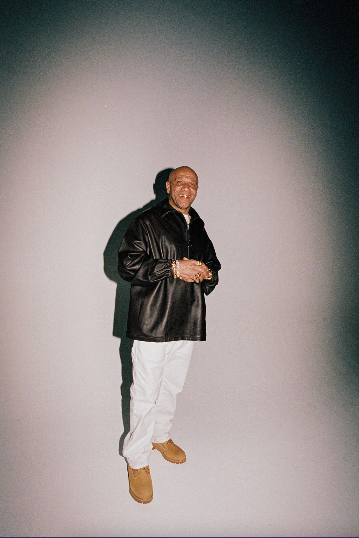 A man with gold teeth smiling, wearing a black leather jacket, white pants, tan boots, and multiple gold jewelry items, standing against a plain gradient background.