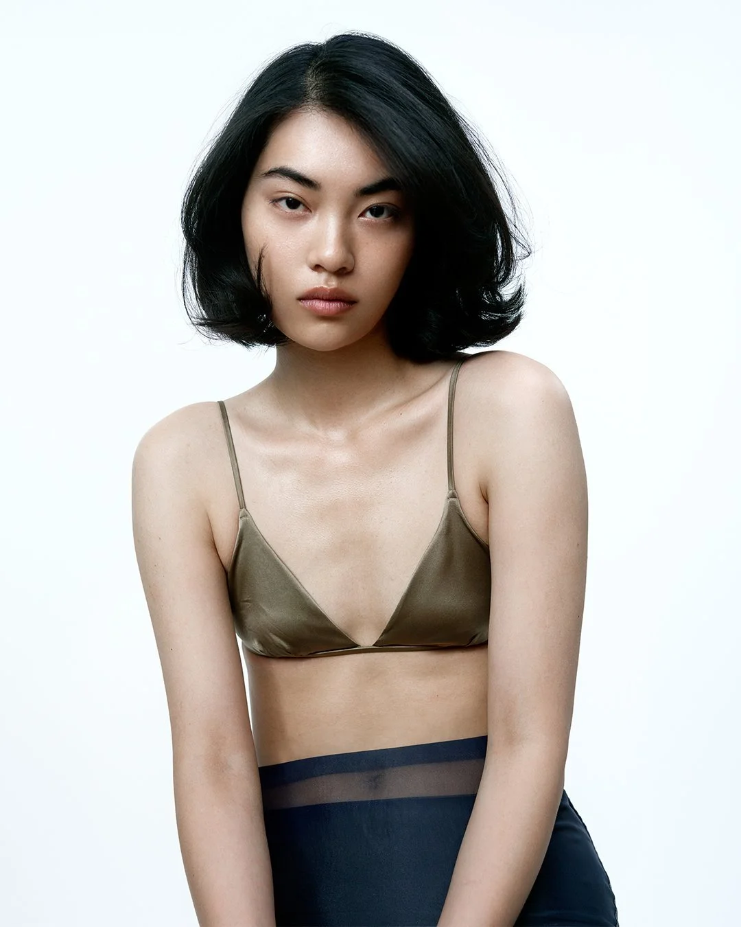 A young woman with black hair and fair skin, wearing a beige spaghetti strap crop top and black high-waisted pants, posing against a plain white background.