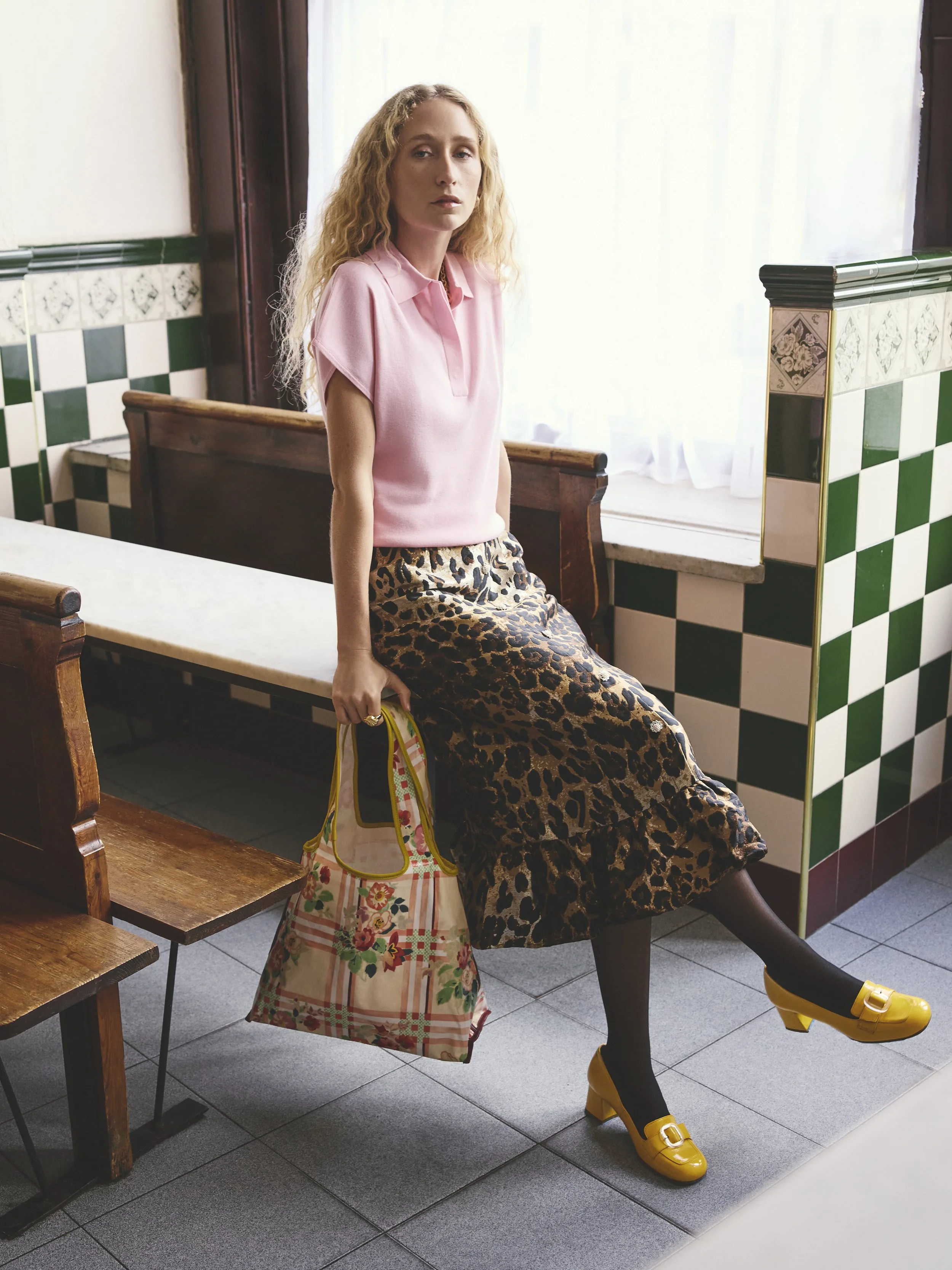 A woman with long, wavy blonde hair sitting on a wooden bench in a bright room, wearing a pink Polo shirt, leopard print skirt, black tights, and yellow shoes, holding a floral-patterned bag.