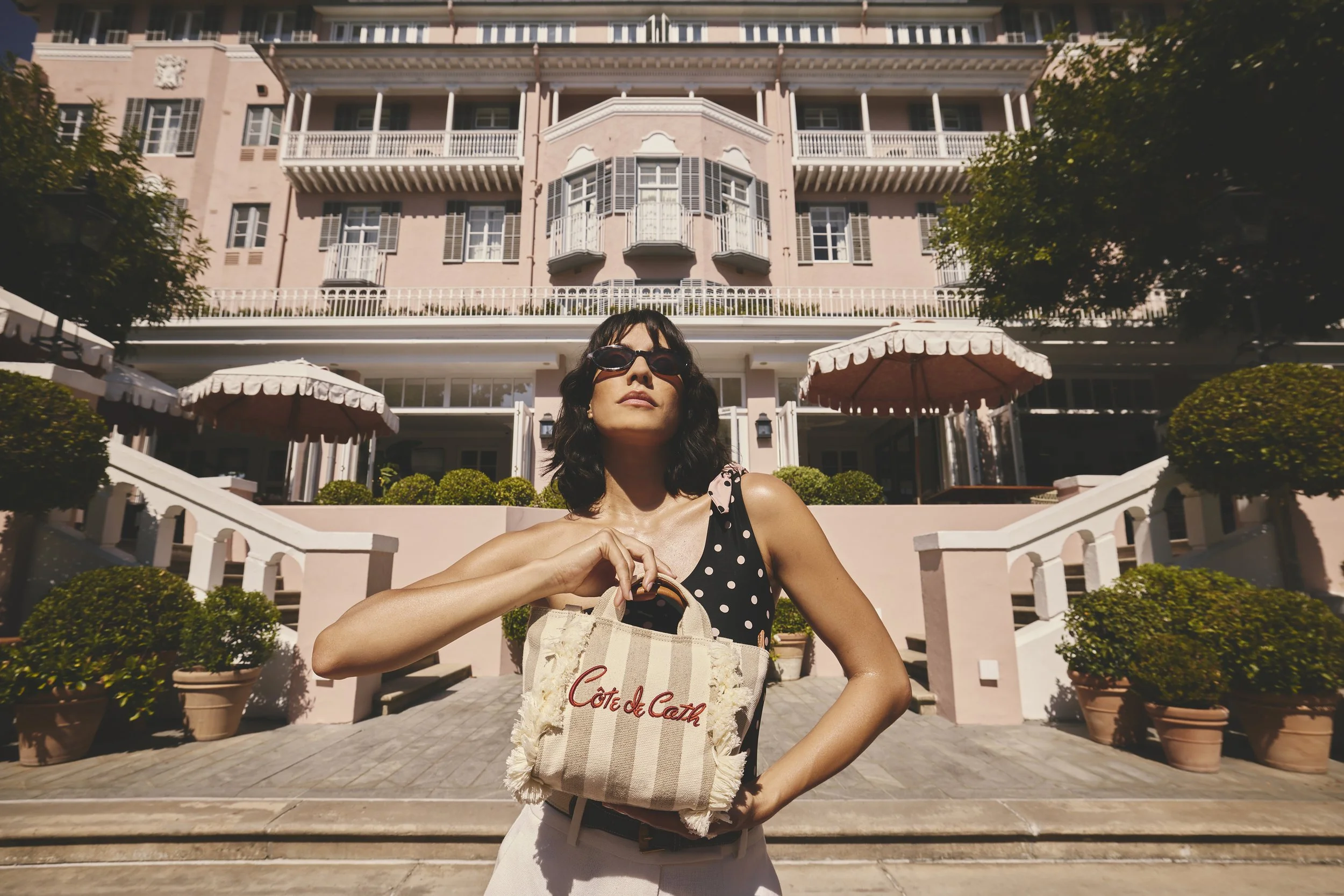 A woman wearing sunglasses and a polka dot top stands in front of a pink multi-story building, holding a beige tote bag with the words 'Côte de Catz' on it. The building has balconies and a stairway leading to the entrance, with potted plants and umb