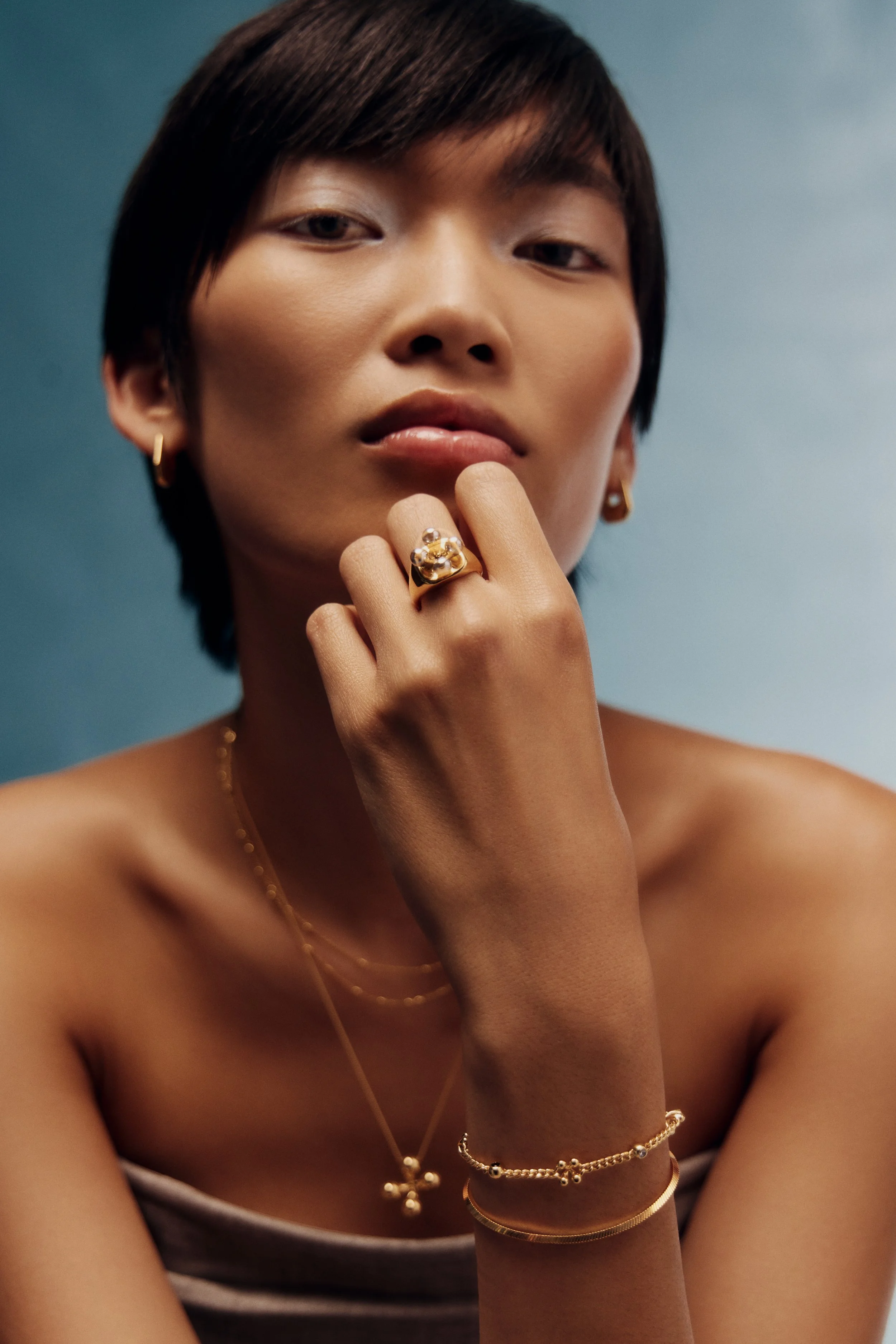 A woman with short dark hair and gold jewelry, touching her lips with her hand, against a blue background.