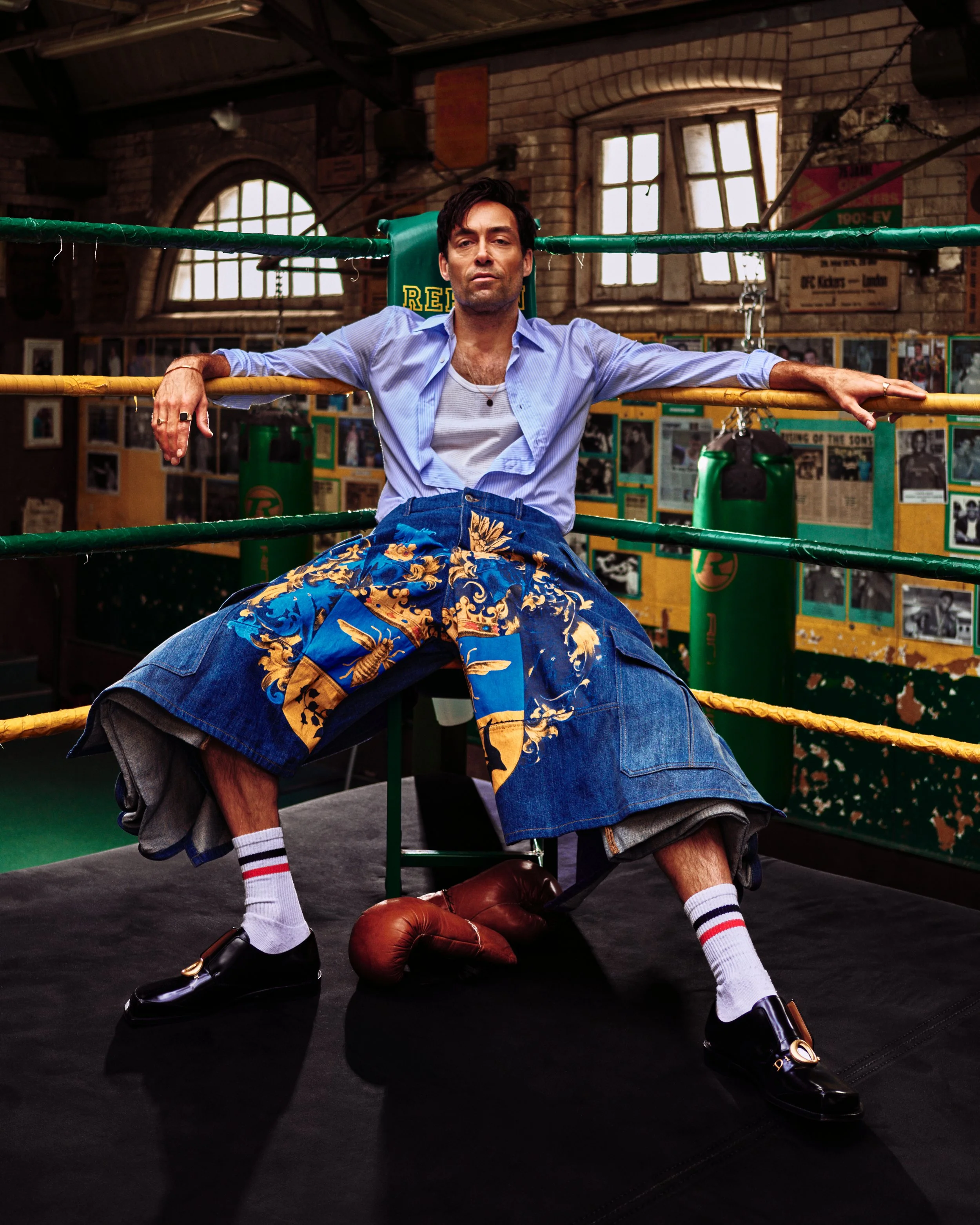 Alexander Hassell the actor in a boxing ring with his arms resting on the ropes, wearing a light purple button-up shirt, patterned shorts, and shiny black shoes. His expression is serious, and he is surrounded by vintage boxing memorabilia 