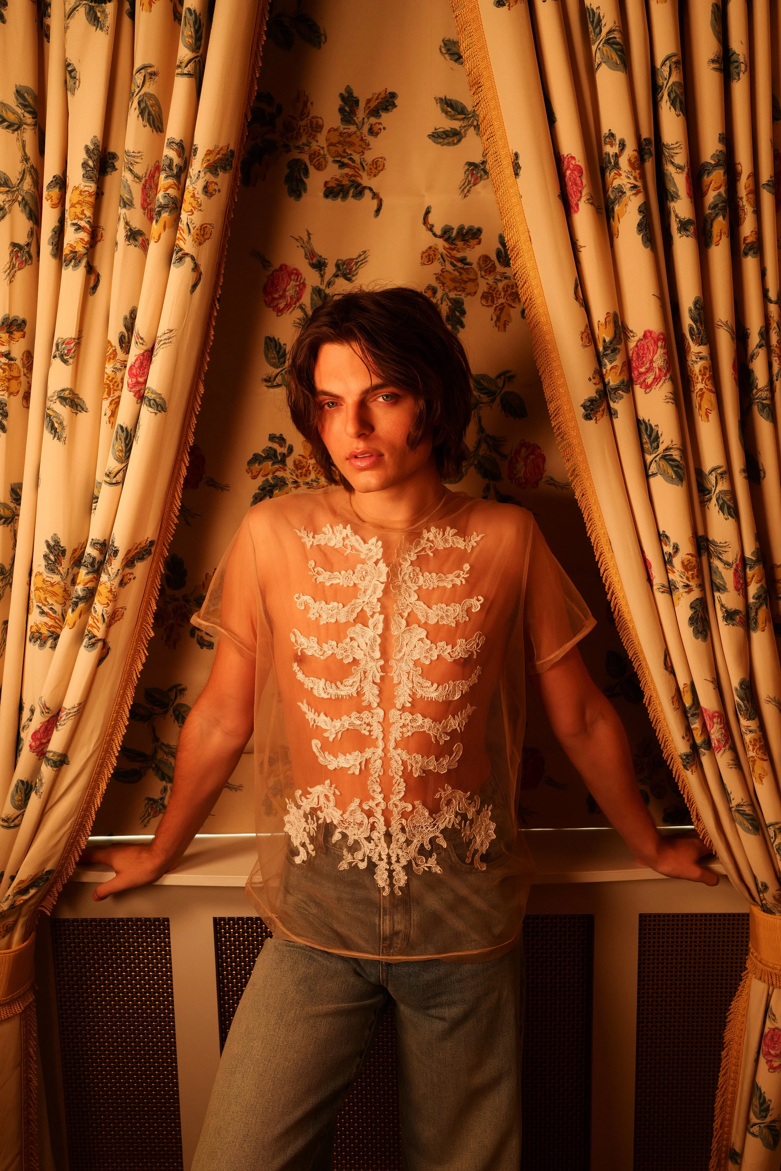 A person with shoulder-length brown hair and light skin, wearing a sheer, peach-colored top with white floral embroidery, standing in a window surrounded by floral-patterned curtains. The person is leaning forward with their arms resting on the windo