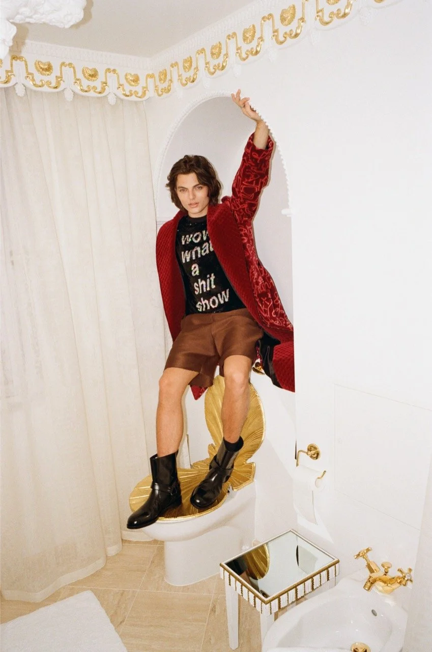 A woman sitting on a decorative gold toilet with a high back, in a room with white walls and gold accents. She is wearing a red coat, a black shirt with white text, brown shorts, and black boots. She has short, wavy hair and is posing with one arm ra