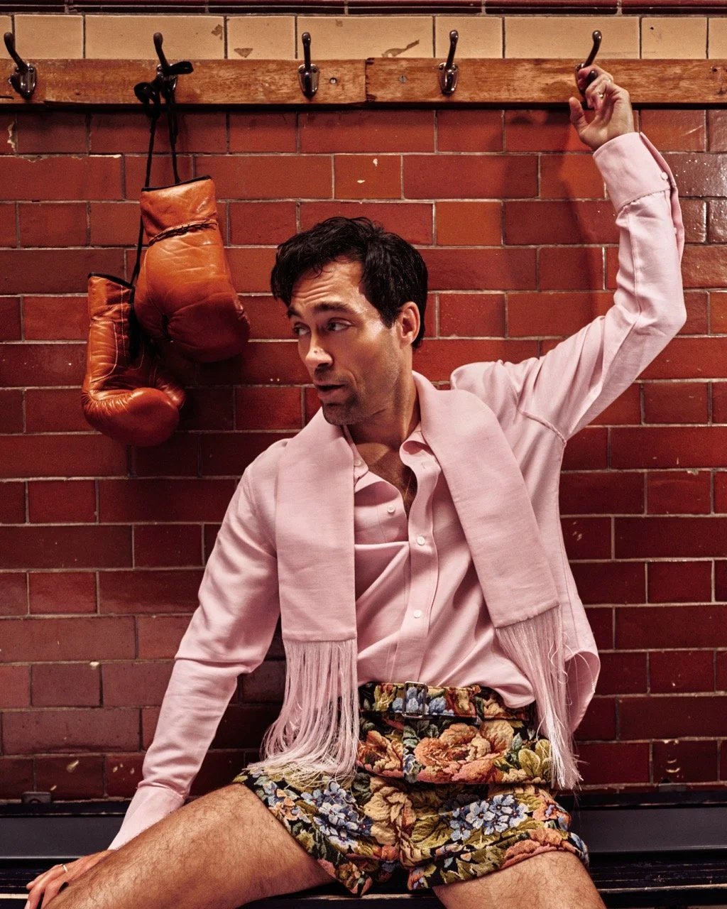 A man sitting against a red brick wall with boxing gloves hanging from hooks above him, wearing a pink shirt and floral shorts.