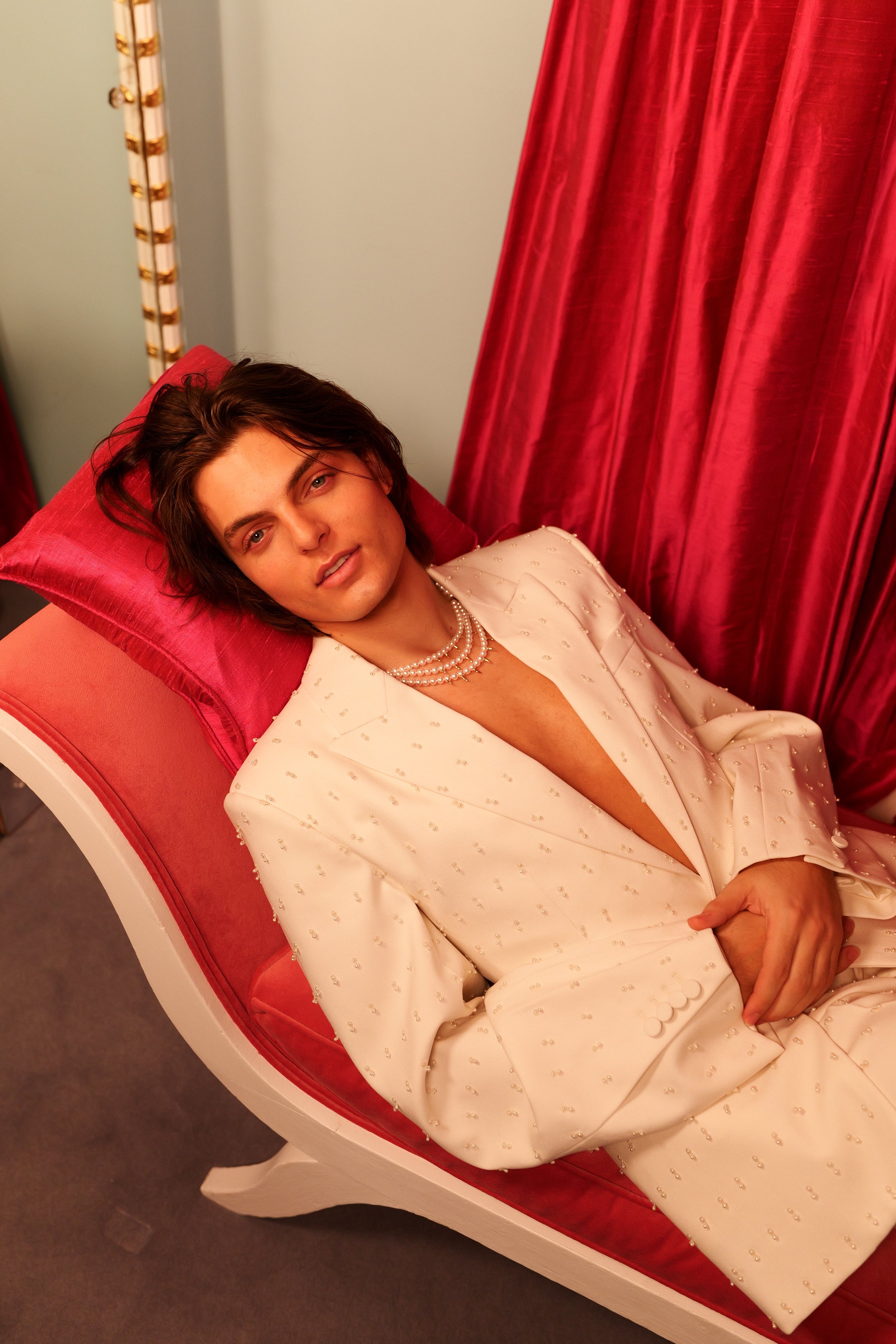 A person with long dark hair lying on a red cushioned lounge chair, wearing a cream-colored blazer with pearl embellishments and a pearl necklace, surrounded by red curtains in the background.
