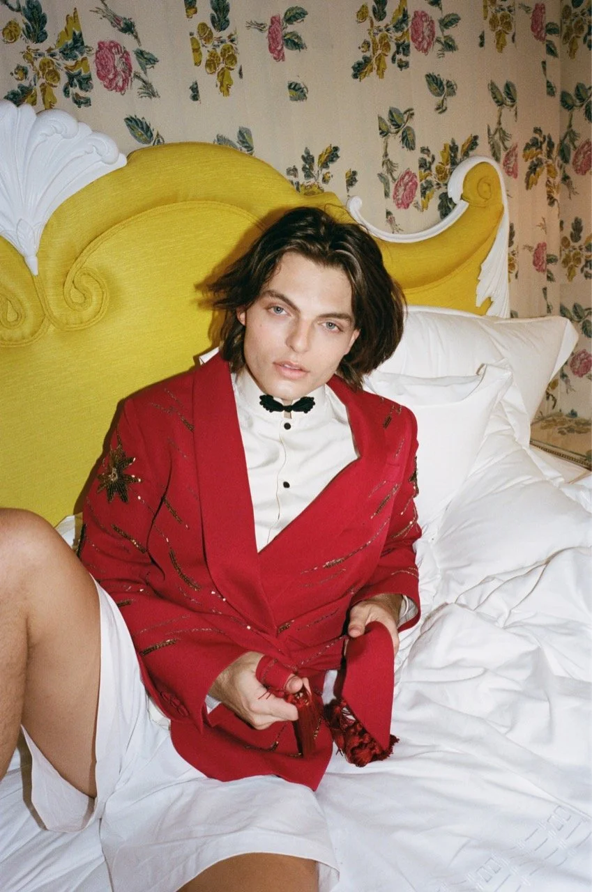 A young man with long dark hair, wearing a white shirt with a bow tie and a red jacket with decorative embroidery, sitting on a bed with white sheets and pillows, in a room with floral wallpaper and a yellow upholstered headboard.