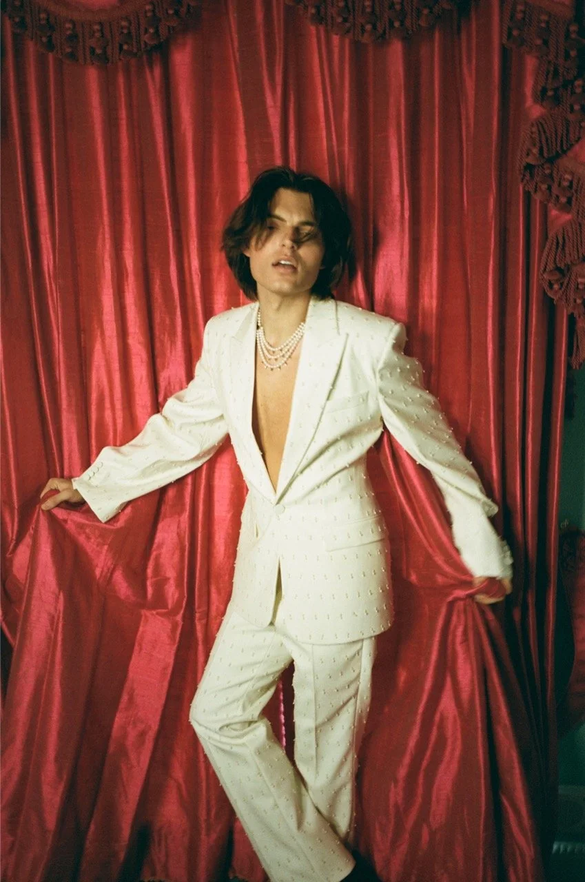 Person in a white suit with pearl necklaces posing in front of a red curtain.