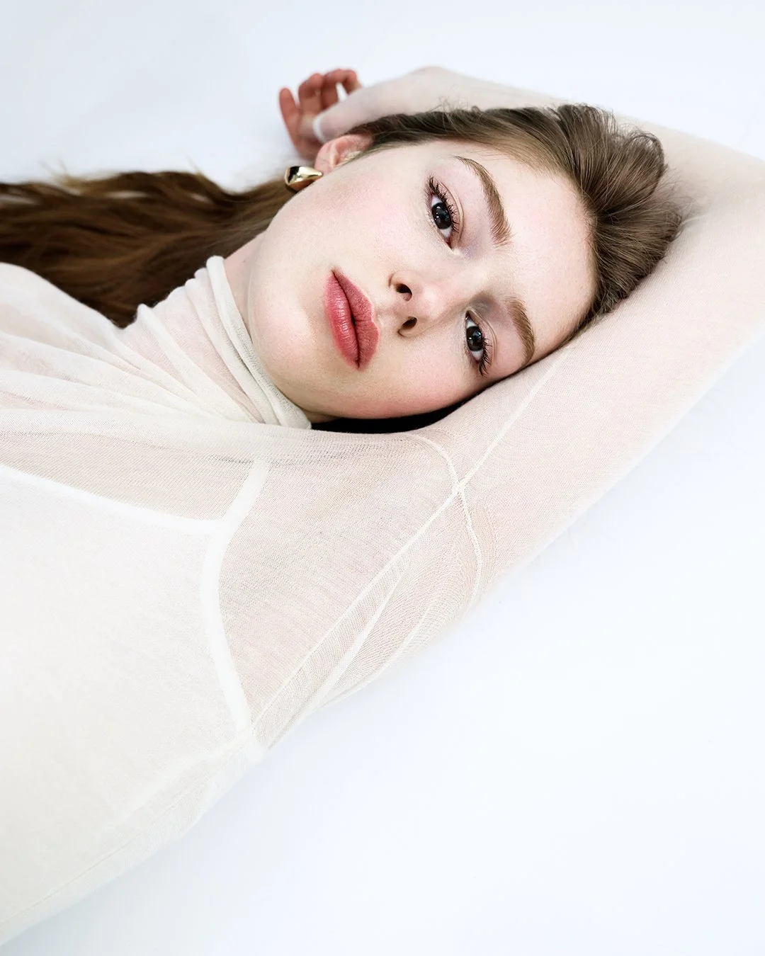 A woman with long brown hair and light skin lying down on a beige pillow, looking at the camera with a relaxed expression.