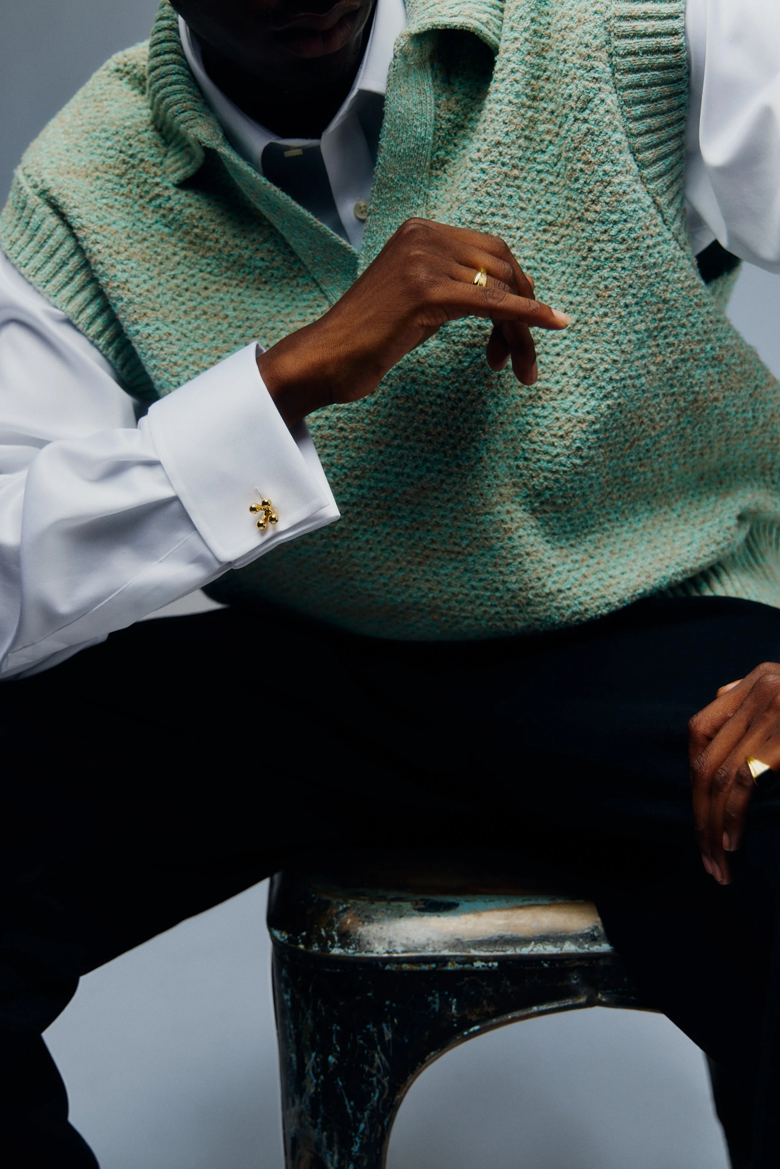 A person wearing a white dress shirt with gold cufflinks and a textured green vest is sitting on a vintage checkered stool. Their right hand, with a gold ring, is resting on their lap, and their left hand, also wearing a ring, is partially visible.