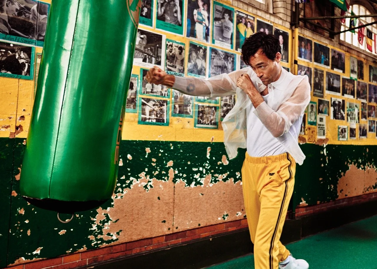 A man in yellow pants and a white shirt practices boxing by punching a green punching bag in a gym with walls decorated with numerous black-and-white photographs.